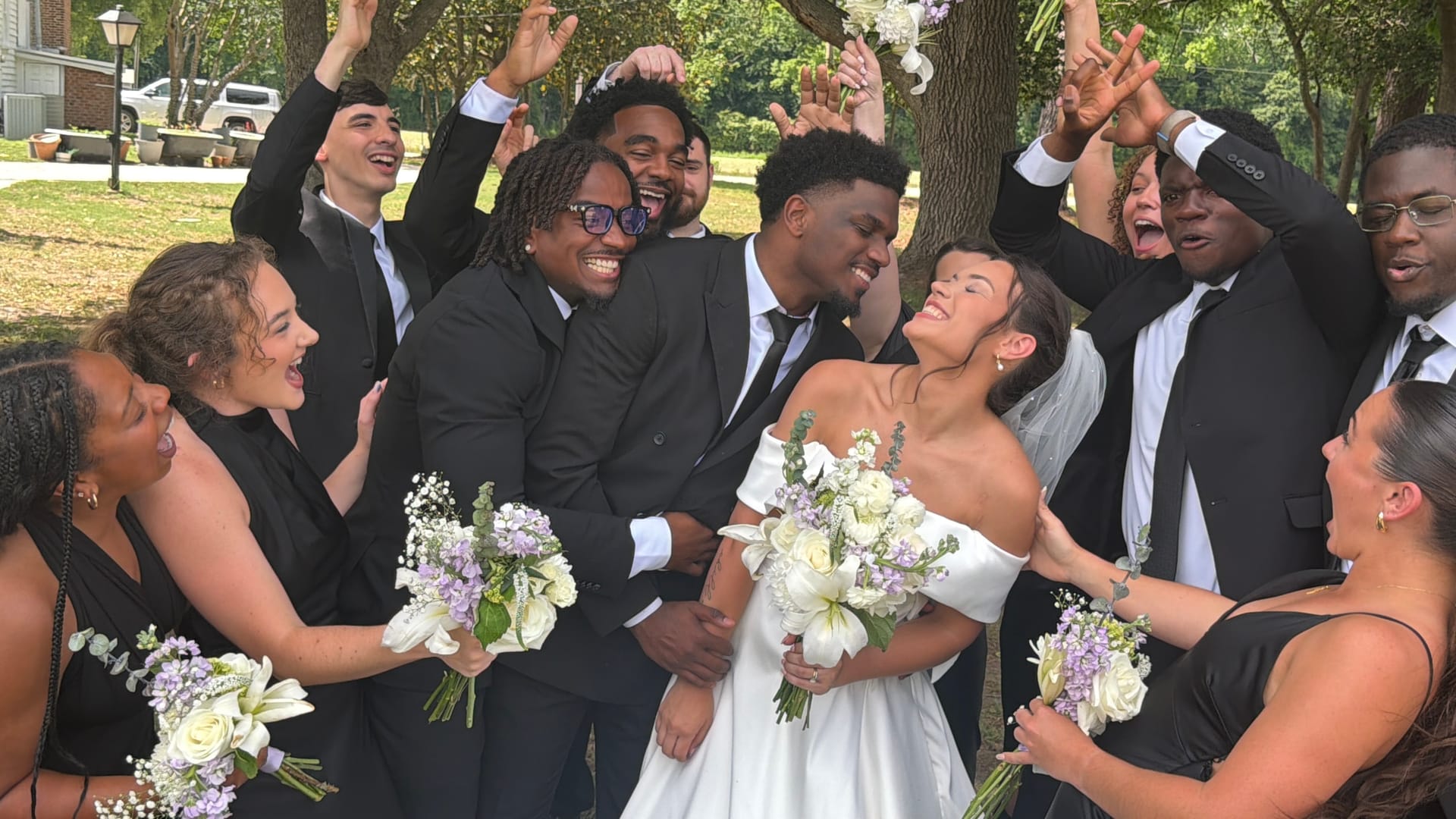 Bride in white off-shoulder gown and groom in black suit share a romantic moment surrounded by cheering bridesmaids and groomsmen holding purple and white bouquets under oak trees.