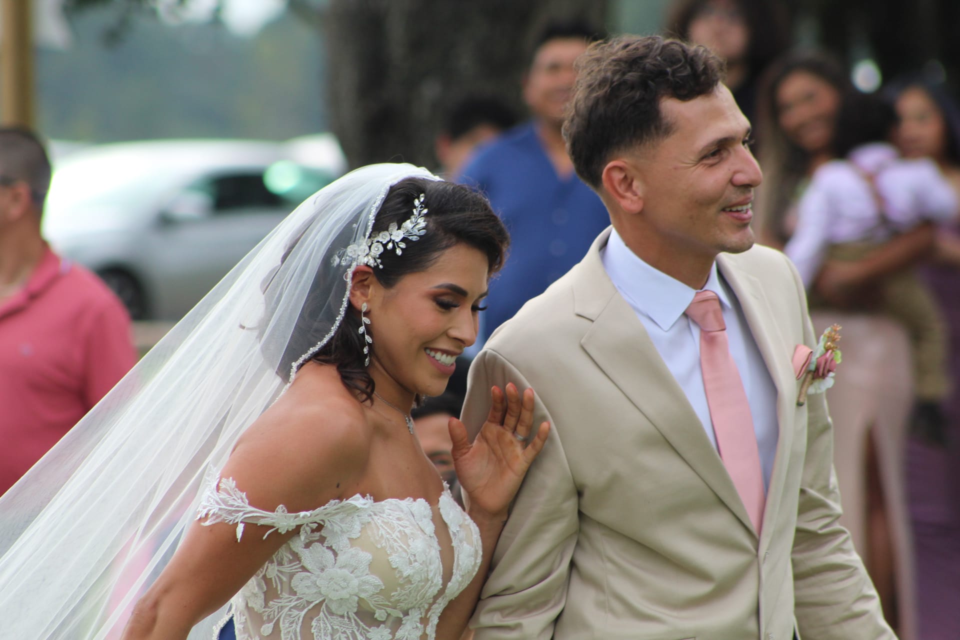 Smiling bride in a lace off-shoulder gown and veil walks arm in arm with groom in a tan suit and pink tie on the ceremony lawn at Scarborough House.