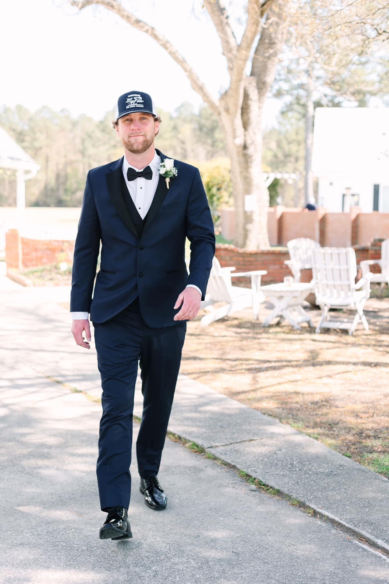 Groom in a navy tuxedo with black bow tie and boutonniere walks confidently down a sunlit pathway at Scarborough House historic estate wedding venue.