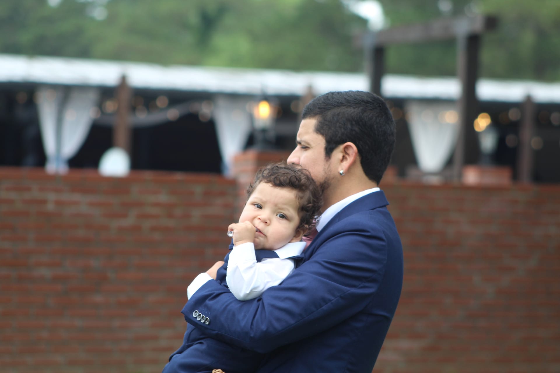 A man in a navy suit tenderly holds a curly-haired toddler dressed in a matching suit at an outdoor wedding venue with a brick wall and draped pavilion in the background.