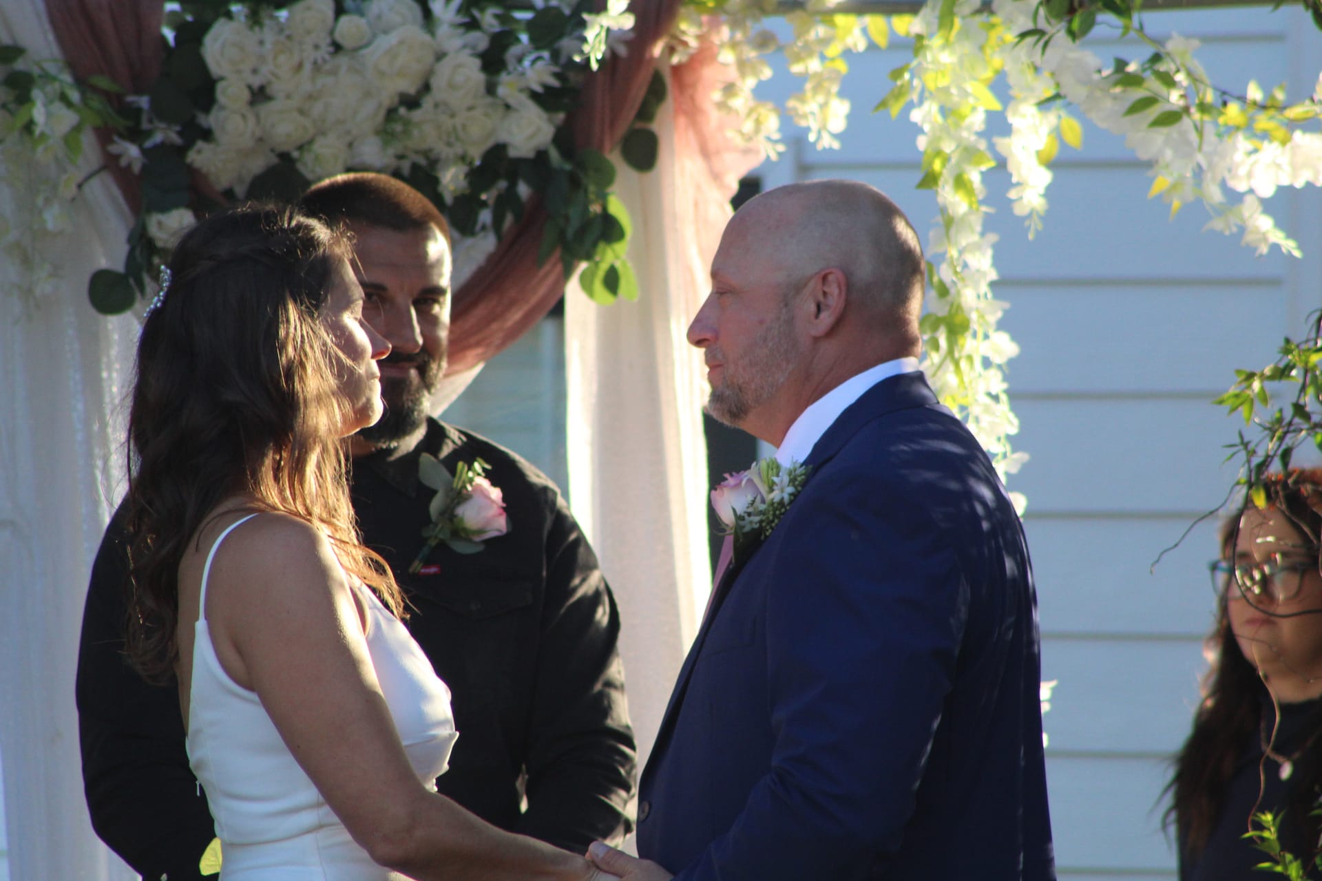 Bride and groom facing each other during outdoor wedding ceremony beneath a white floral and greenery arch at Scarborough House historic estate.