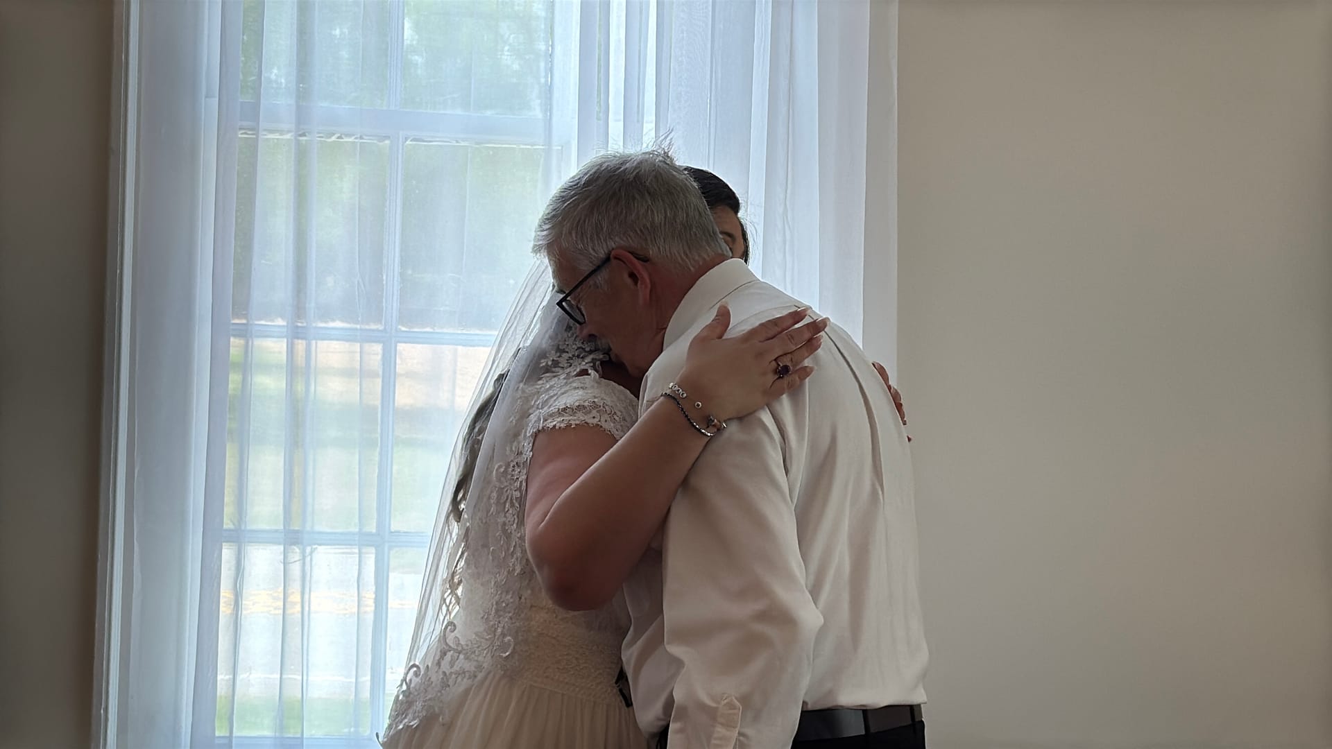 A bride in a lace gown and veil shares a heartfelt embrace with an older gentleman in a white dress shirt in front of a sunlit window at Scarborough House.