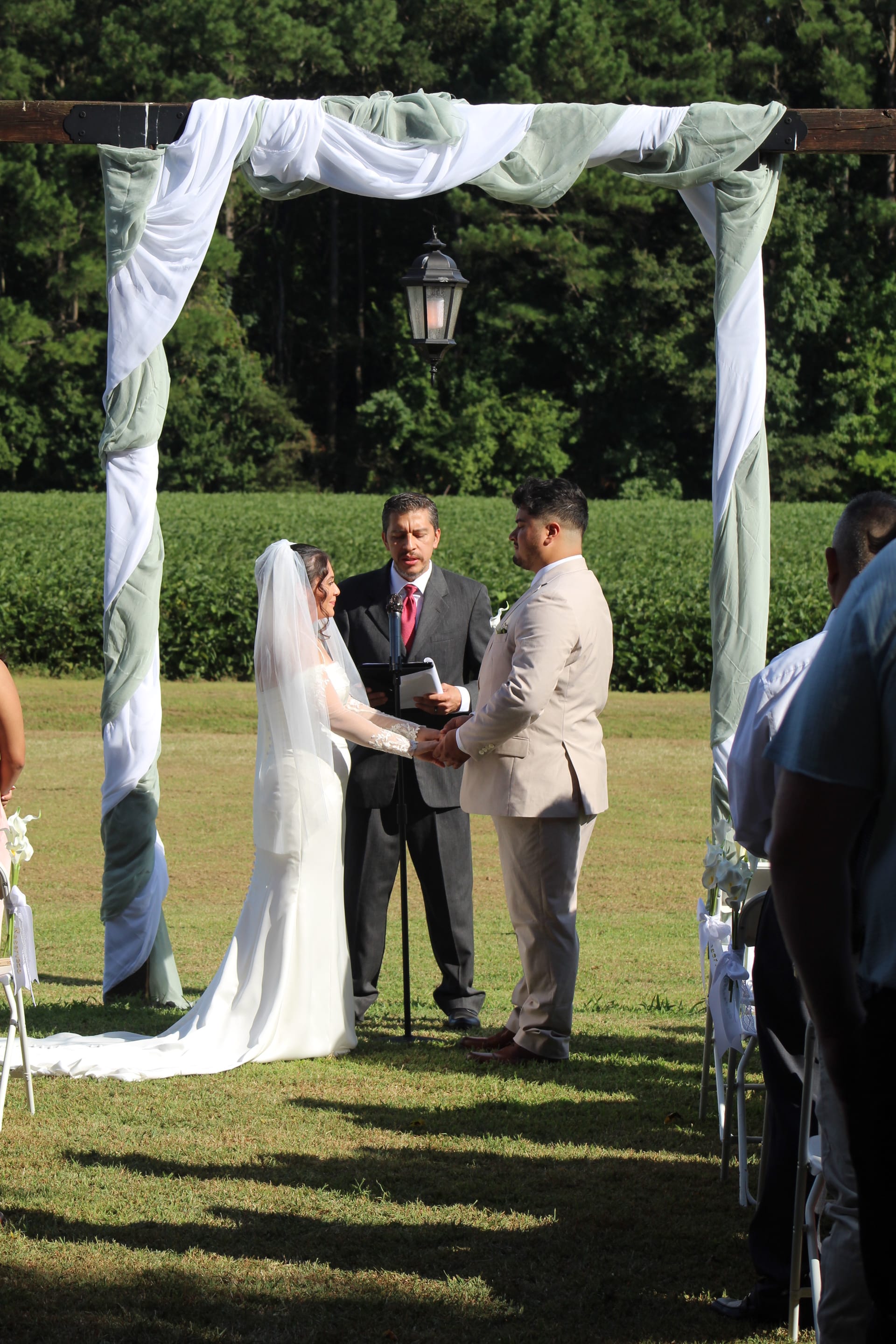 Bride in white gown and veil exchanges vows with groom in tan suit under a draped wooden arch on the ceremony lawn at Scarborough House, with green farmland fields and trees in the background.
