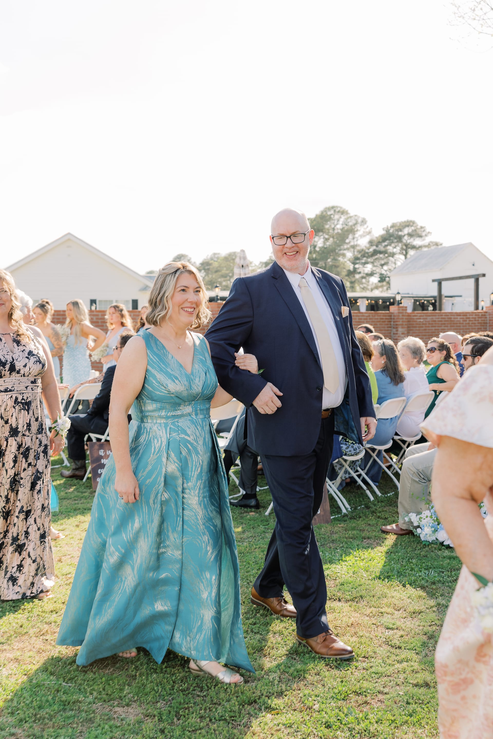 A smiling woman in a teal gown walks arm-in-arm with a man in a navy suit down the outdoor ceremony aisle at Scarborough House.