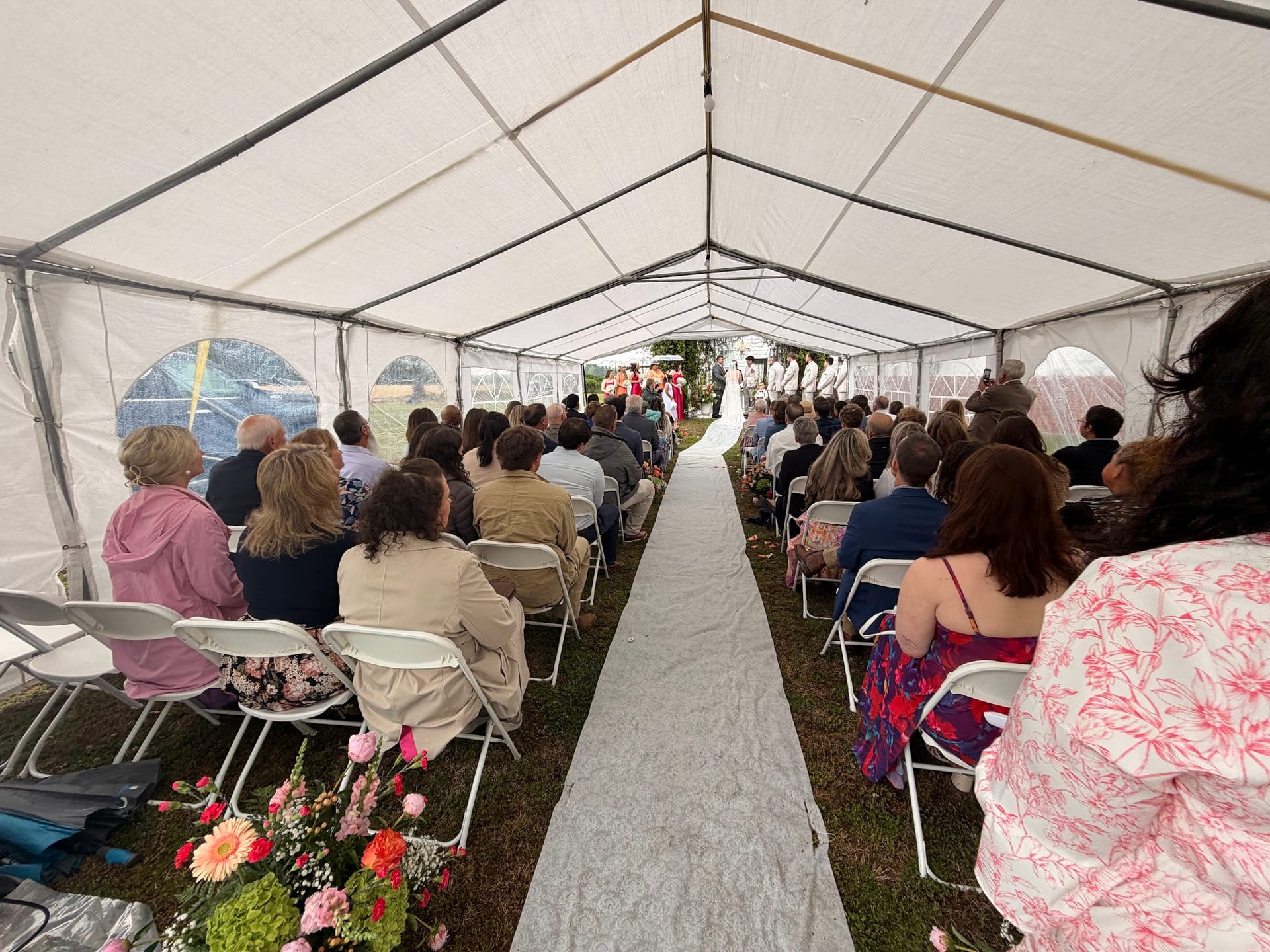 Guests seated in white folding chairs under a large marquee tent watch a wedding ceremony at the end of a white aisle runner lined with pink floral arrangements at Scarborough House.