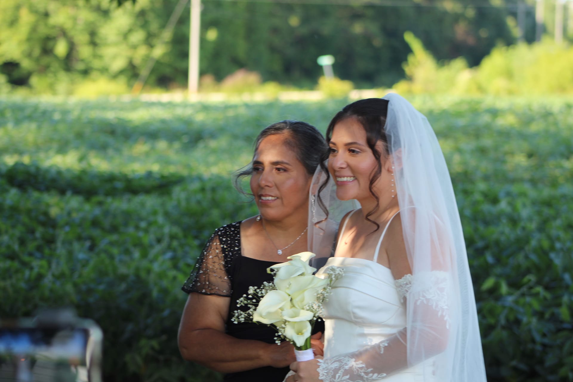 A smiling bride in a white gown and veil holds a bouquet of white calla lilies while standing close to an older woman in a black dress against a lush green garden backdrop at Scarborough House.