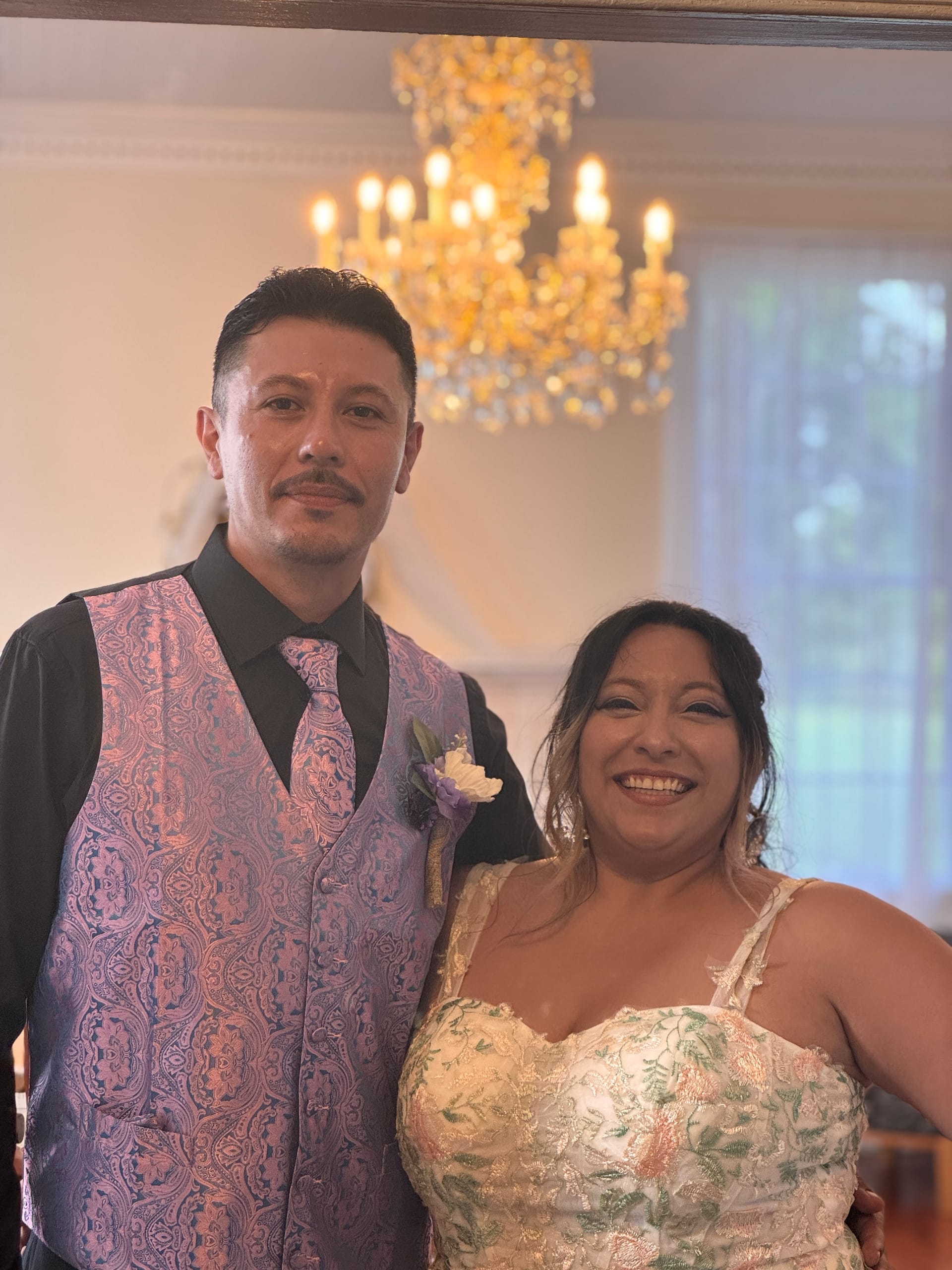 Bride in a floral embroidered gown and groom in a paisley vest and boutonniere smile together beneath a golden chandelier inside Scarborough House historic estate.