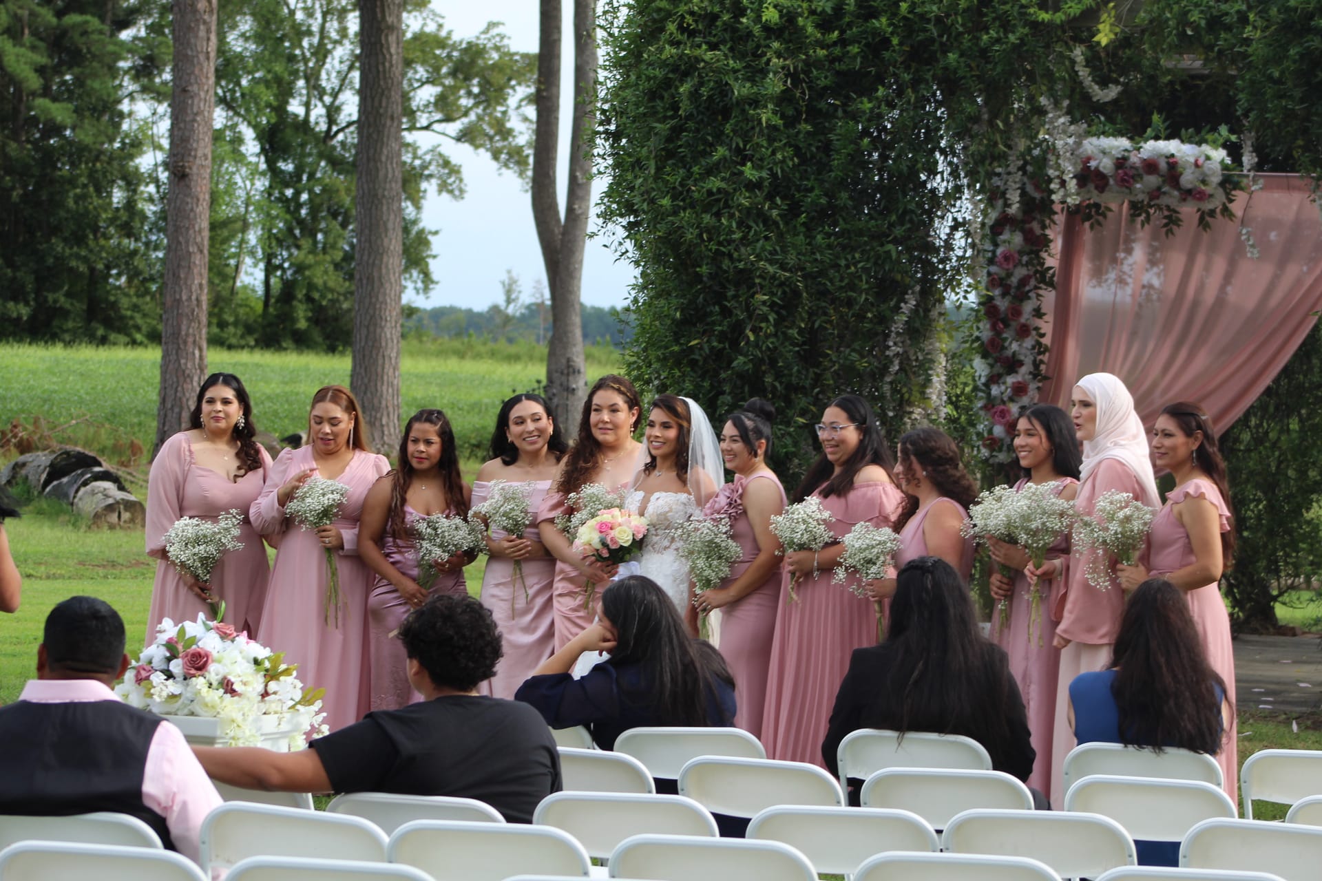 A bride in a white gown and veil stands surrounded by bridesmaids in dusty-rose dresses holding baby's breath bouquets on the outdoor ceremony lawn at Scarborough House, with a floral-draped arch and rolling fields in the background.