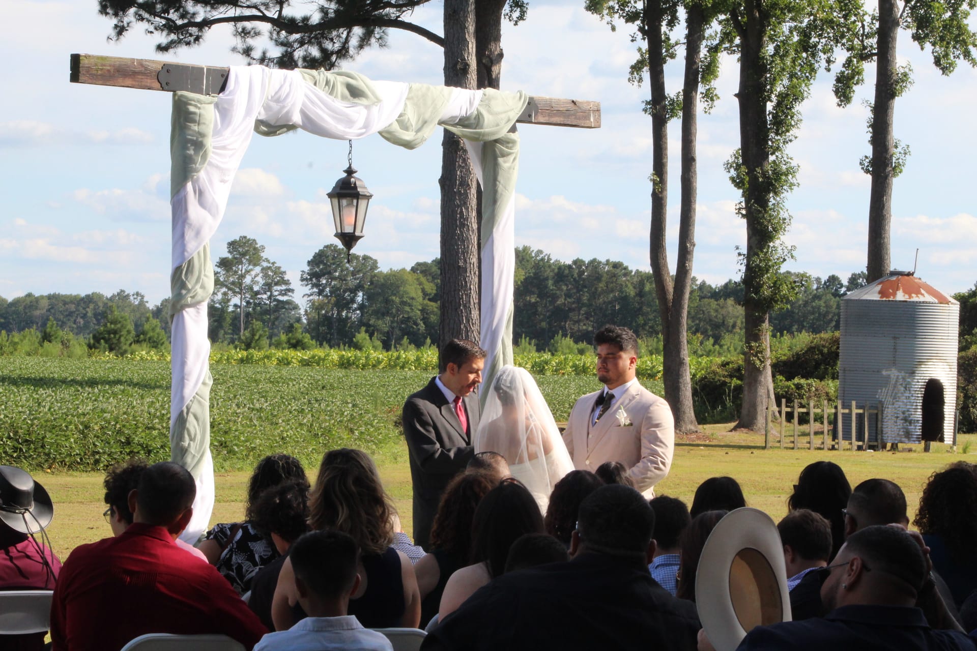 Outdoor wedding ceremony at Scarborough House with a couple exchanging vows beneath a rustic wooden arch draped in white and sage fabric, surrounded by seated guests with open farmland fields and tall pines in the background.