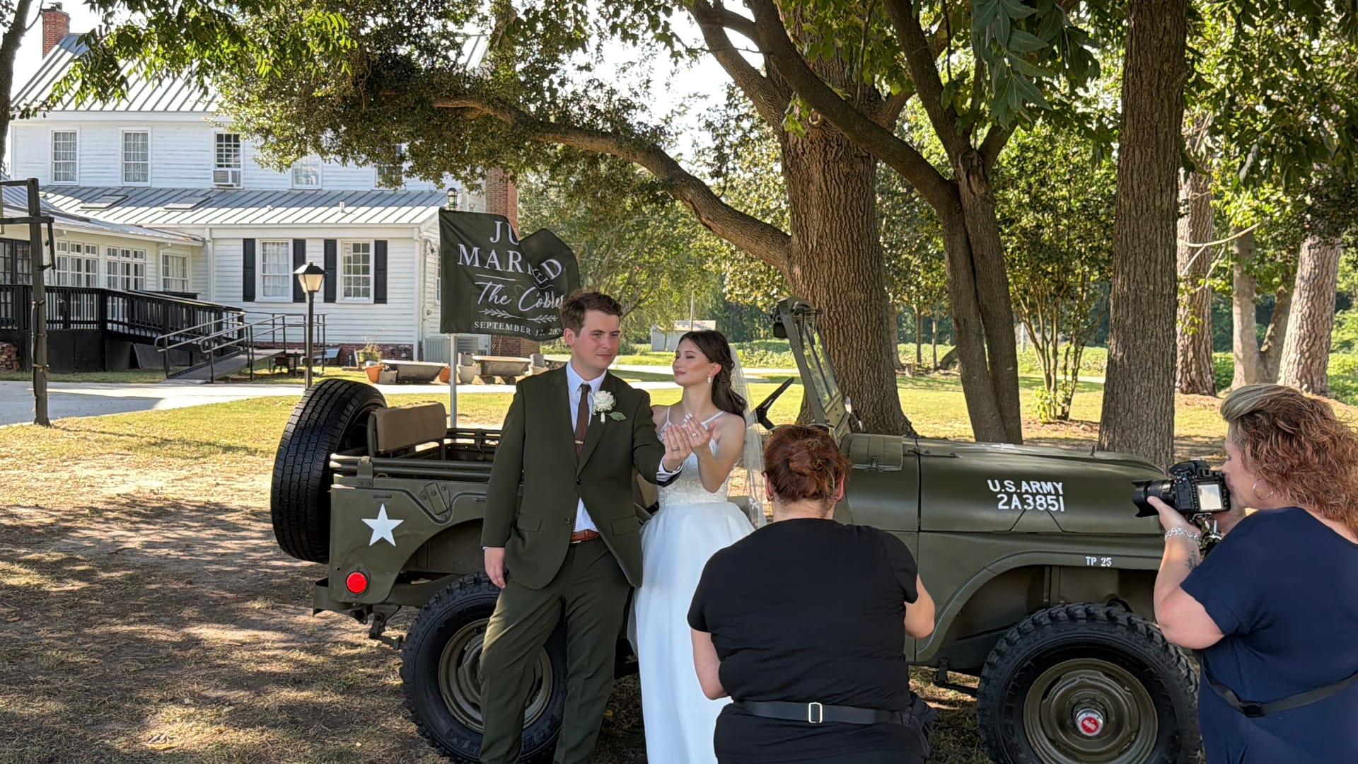 Newlyweds pose in front of a vintage U.S. Army Jeep adorned with a Just Married banner beneath oak trees at Scarborough House historic estate wedding venue.