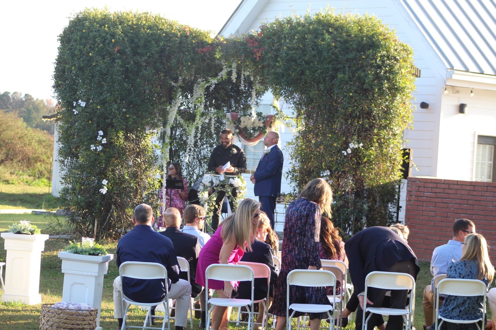 Guests seated on white folding chairs watch an outdoor wedding ceremony beneath a lush ivy-covered arch with white floral accents in front of a historic white farmhouse at Scarborough House.