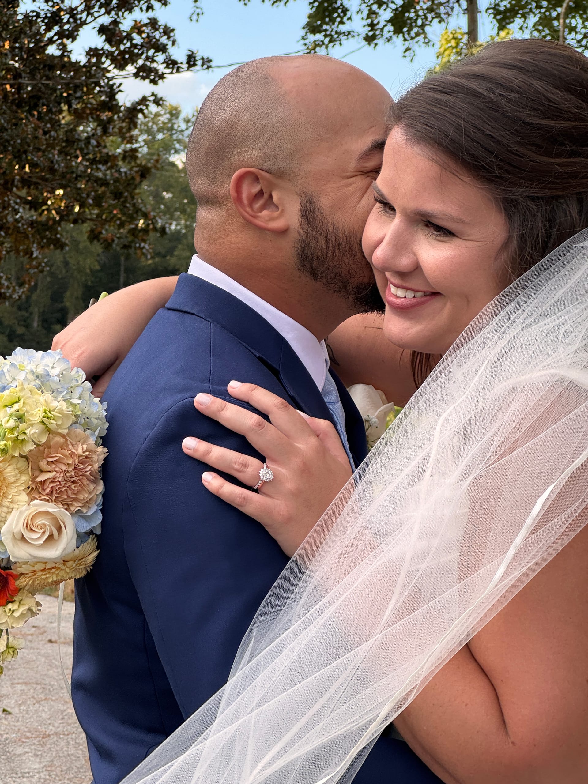 Bride smiling joyfully as groom kisses her cheek outdoors at Scarborough House, her diamond ring and colorful bridal bouquet of hydrangeas and roses prominently displayed.