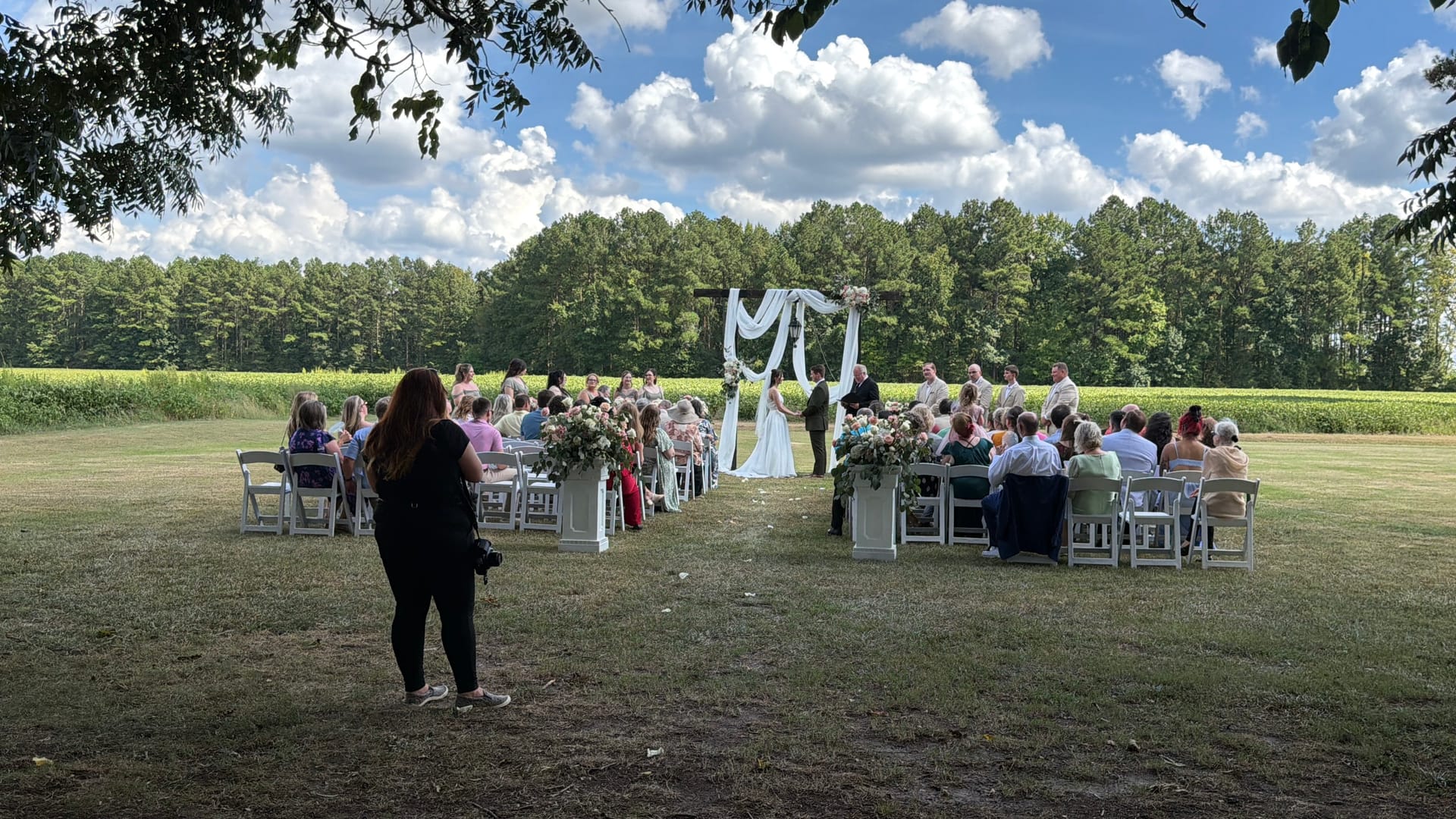 Outdoor wedding ceremony on the Scarborough House ceremony lawn with guests seated in white chairs along a floral-lined aisle leading to a draped wooden arch, surrounded by open farmland fields and tall trees under a bright blue sky.