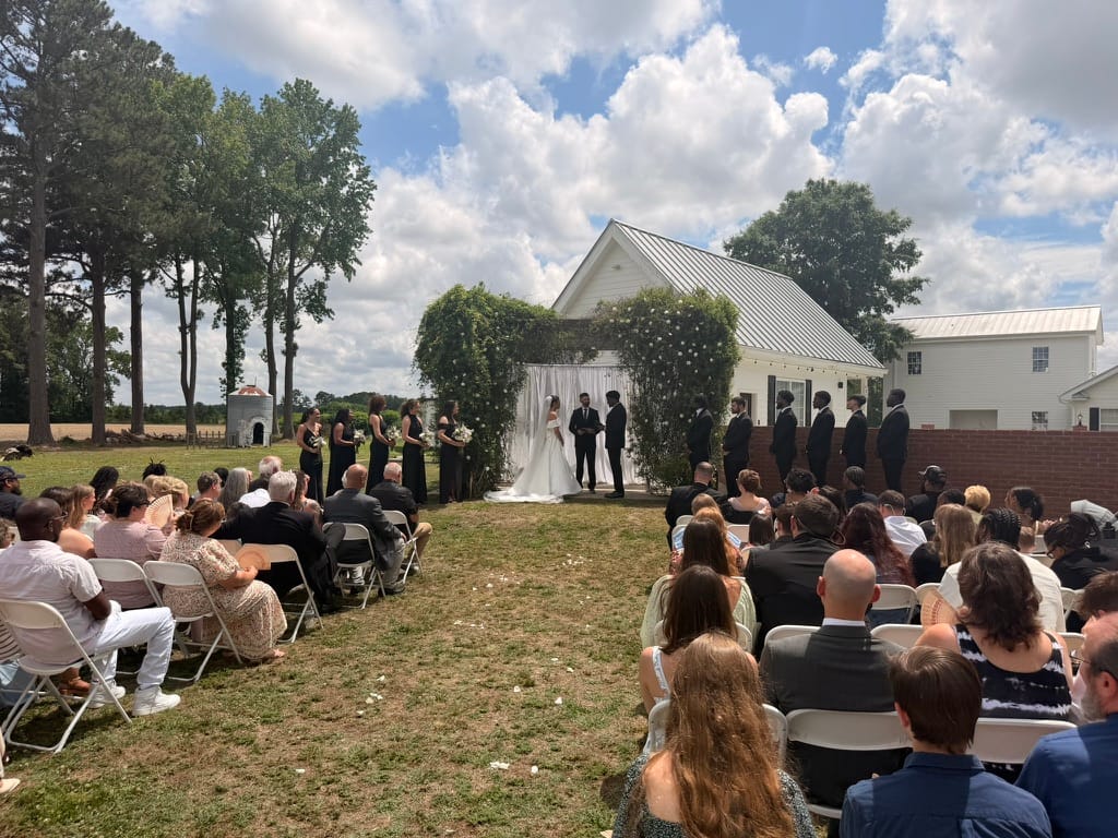 Outdoor wedding ceremony on the ceremony lawn at Scarborough House with guests seated in white chairs, the couple exchanging vows beneath a floral and greenery arch in front of the white historic building.