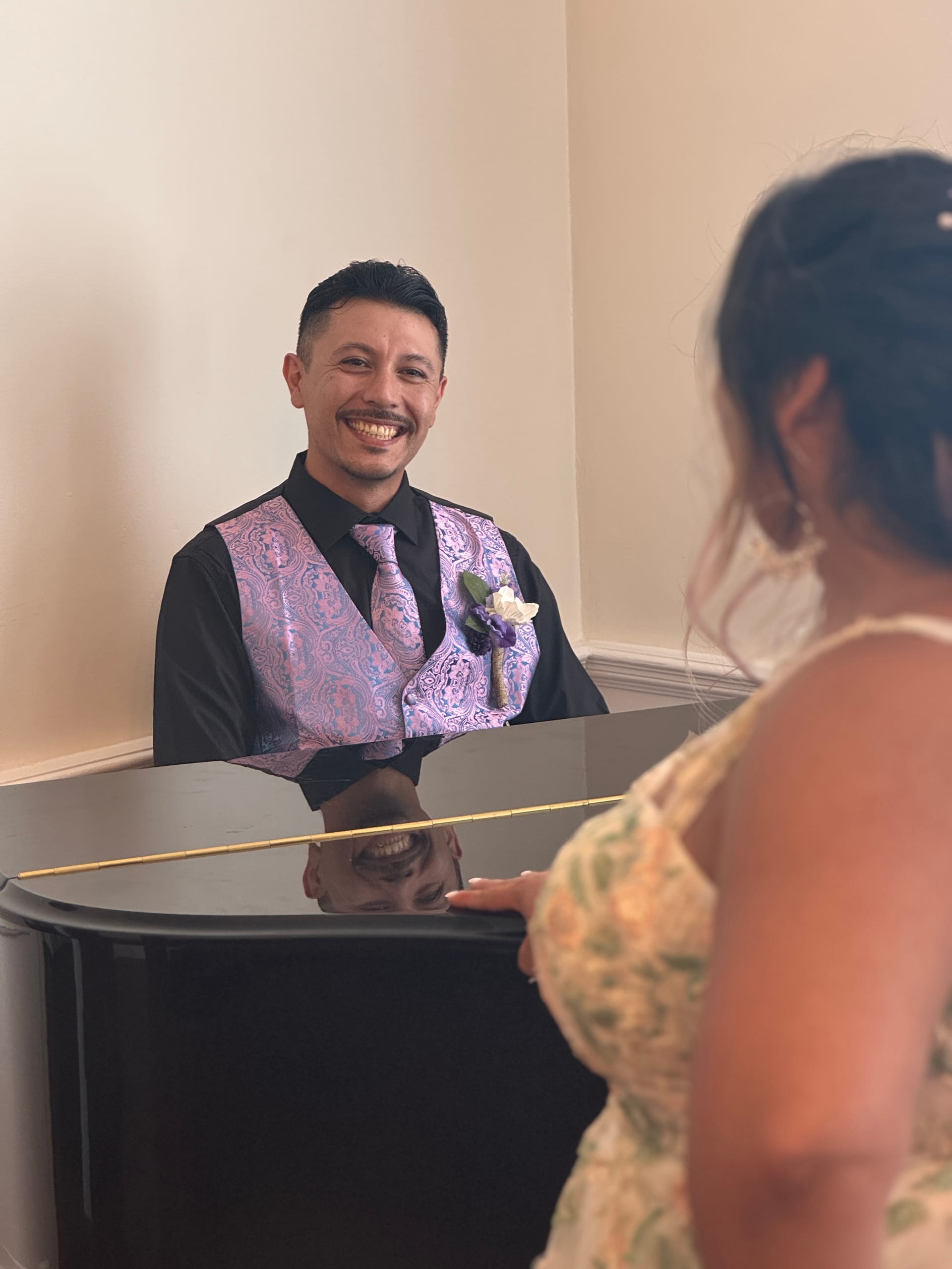 A smiling groom in a purple paisley vest and tie leans over a black grand piano in the grand hall, his reflection visible in the polished lid as a guest looks on.