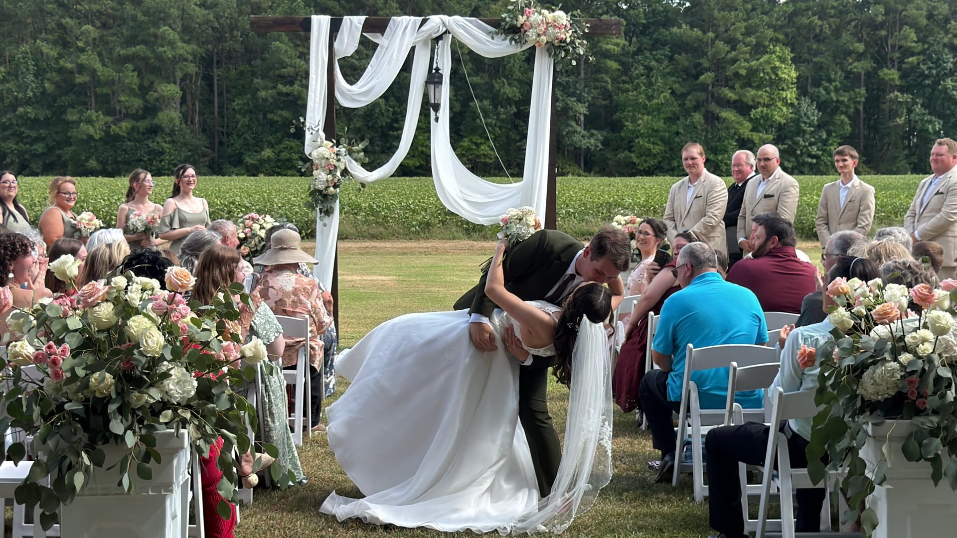 Groom dips bride for a kiss beneath a white-draped wooden arch adorned with blush floral arrangements on the ceremony lawn at Scarborough House, with the wedding party and seated guests looking on against a backdrop of lush green farmland fields.