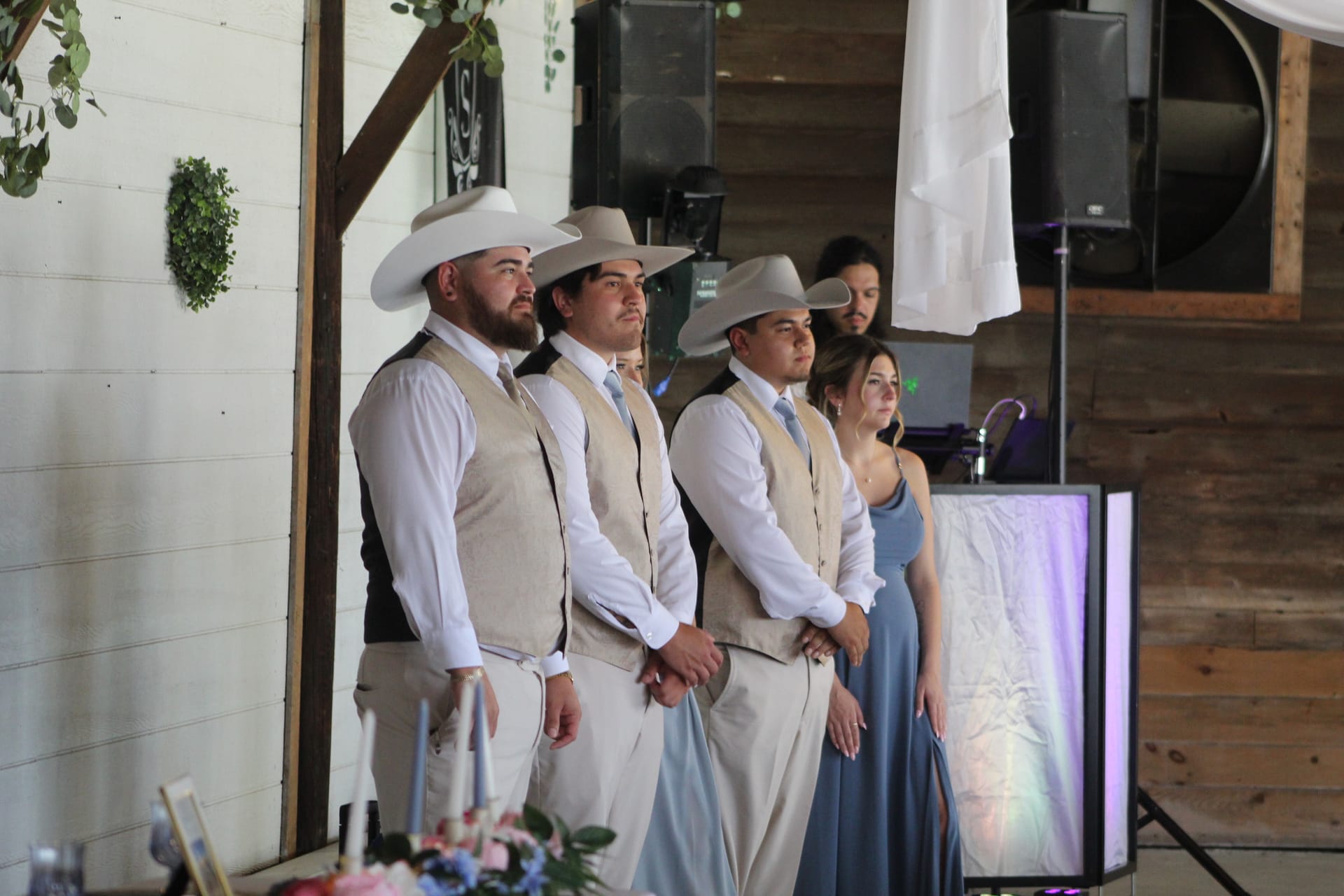 Three groomsmen in tan vests and white cowboy hats stand at attention alongside a bridesmaid in a dusty blue gown during a rustic western-style wedding ceremony inside a barn venue.