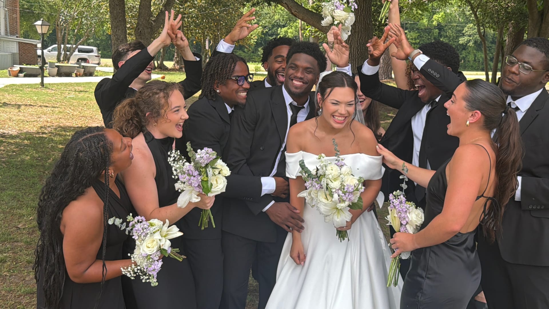 Bride in off-shoulder white gown and groom in black suit laugh joyfully surrounded by their cheering wedding party under oak trees at Scarborough House estate.