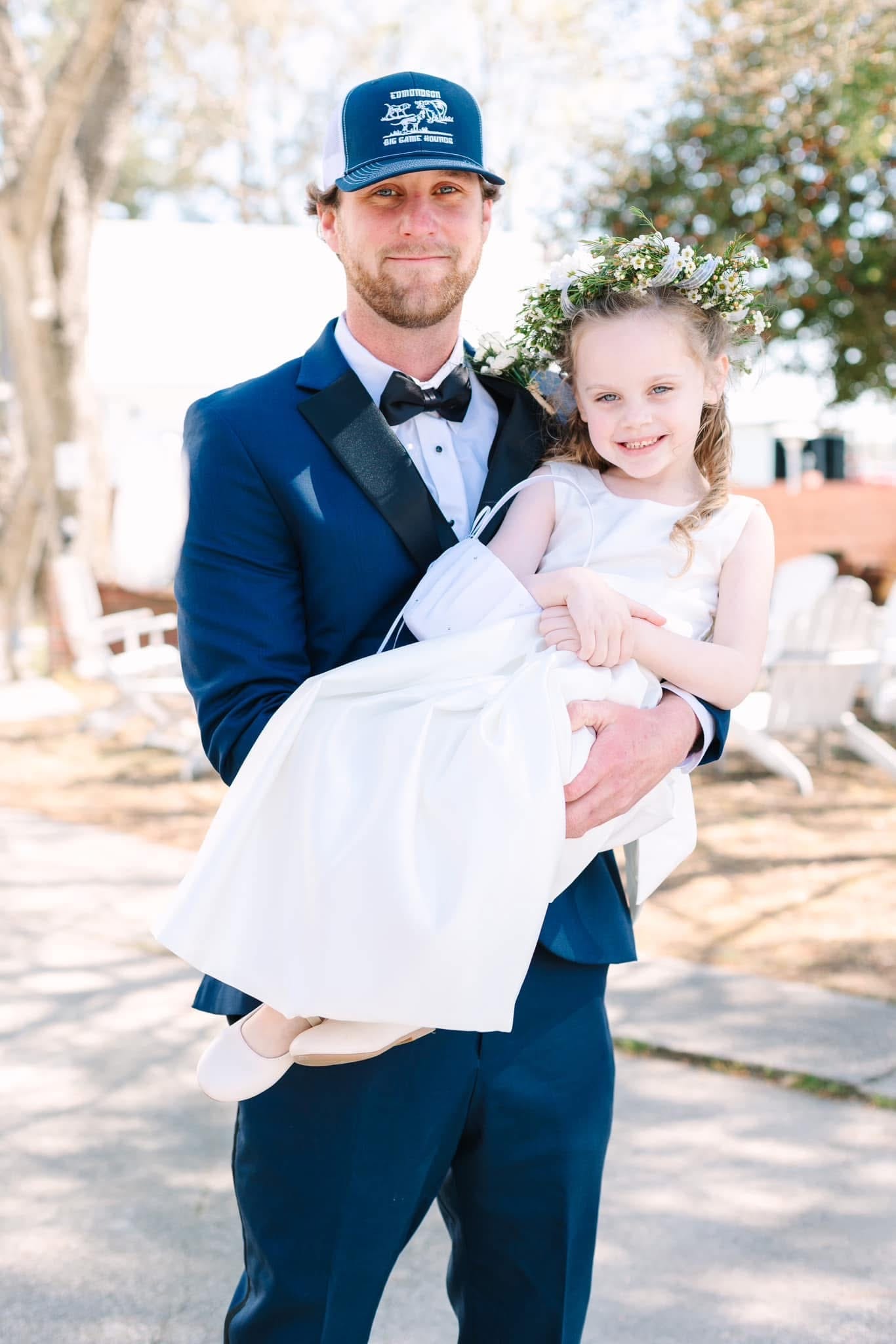 Groom in a navy tuxedo and trucker cap holds a smiling flower girl wearing a white dress and floral crown on the sunlit grounds of Scarborough House estate.
