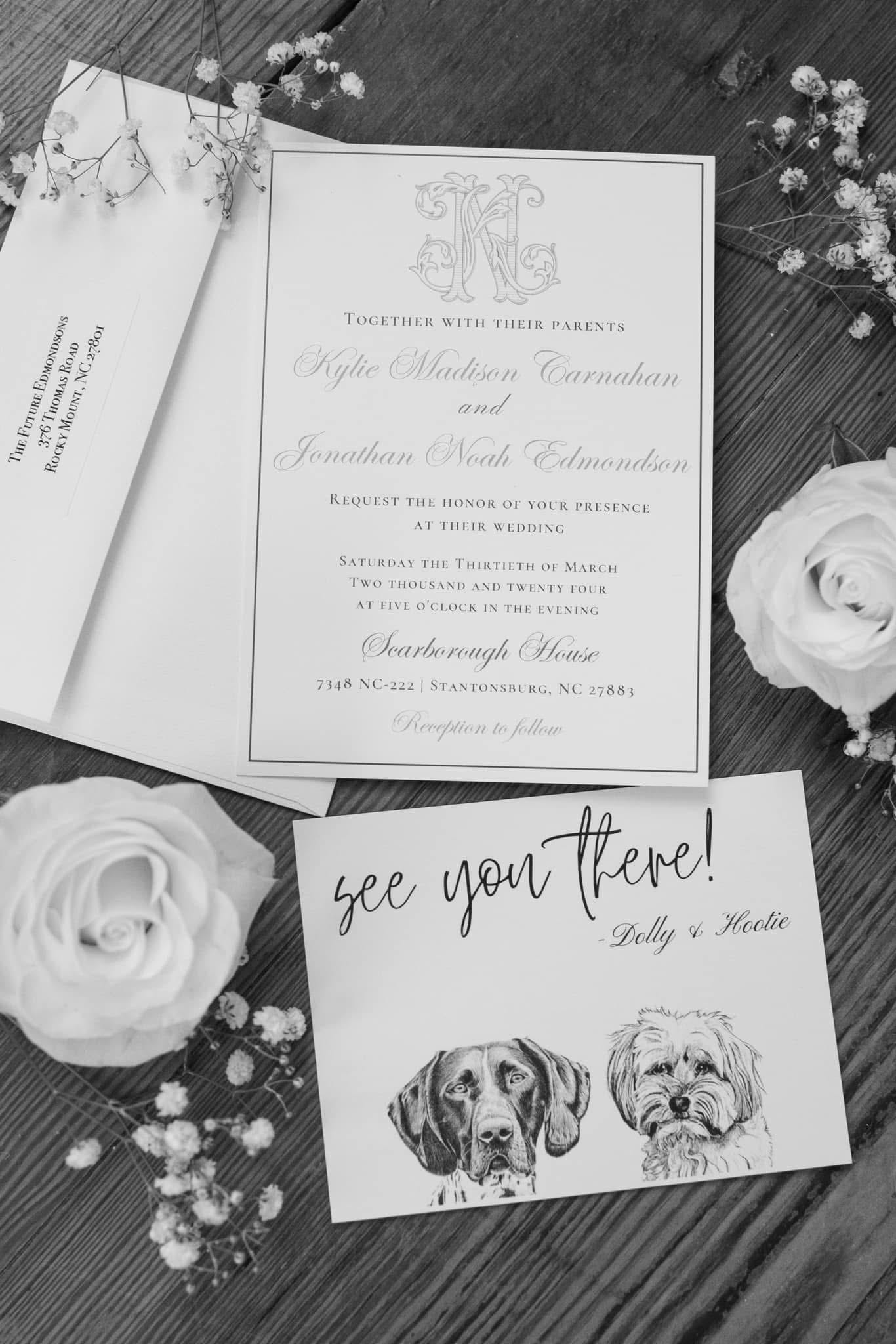 Black and white flat-lay of a formal wedding invitation for Kylie Madison Carnahan and Jonathan Noah Edmondson at Scarborough House, styled with white roses and baby's breath alongside a whimsical dog portrait enclosure card signed by Dolly and Hootie.