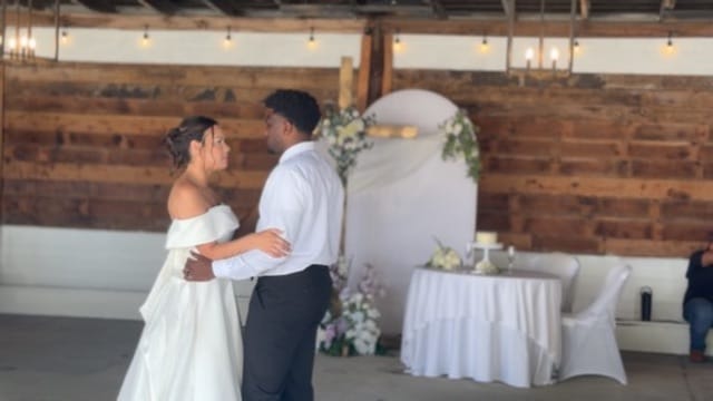 Bride in off-shoulder white gown and groom in white shirt share their first dance in the rustic grand hall adorned with string lights and a floral arch at Scarborough House.