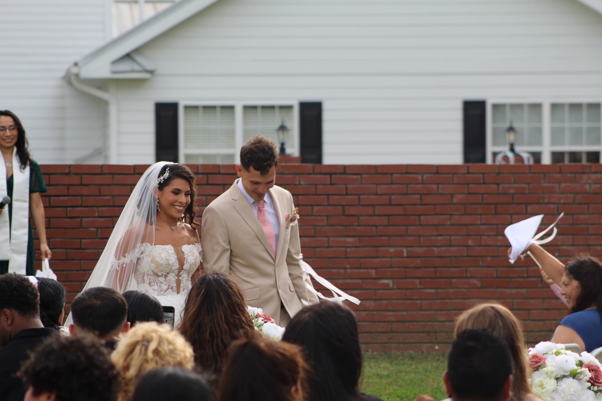 Smiling bride in lace gown and veil walks the recessional with her groom in a tan suit and pink tie past cheering guests on the ceremony lawn at Scarborough House historic estate.