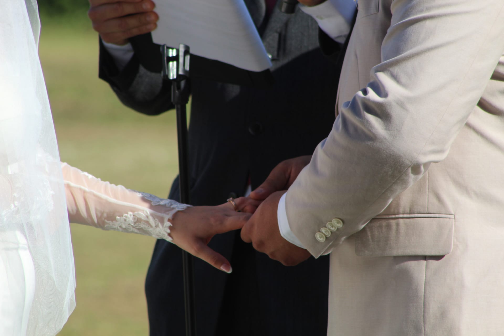Groom in a light gray suit placing a wedding ring on the brides lace-sleeved hand during an outdoor ceremony at Scarborough House.