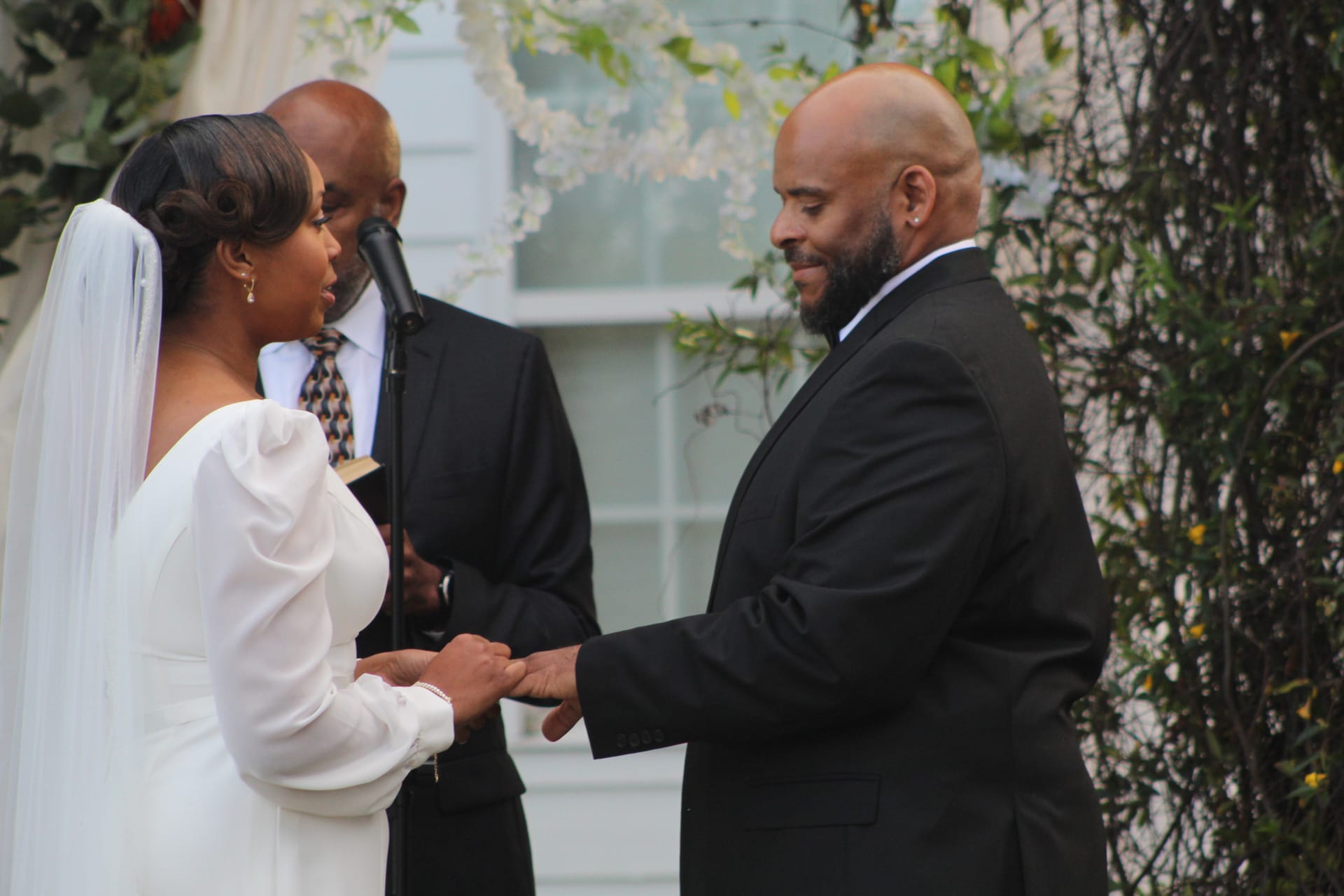 Bride and groom exchange rings during an outdoor wedding ceremony at Scarborough House, framed by a floral arch and officiant.