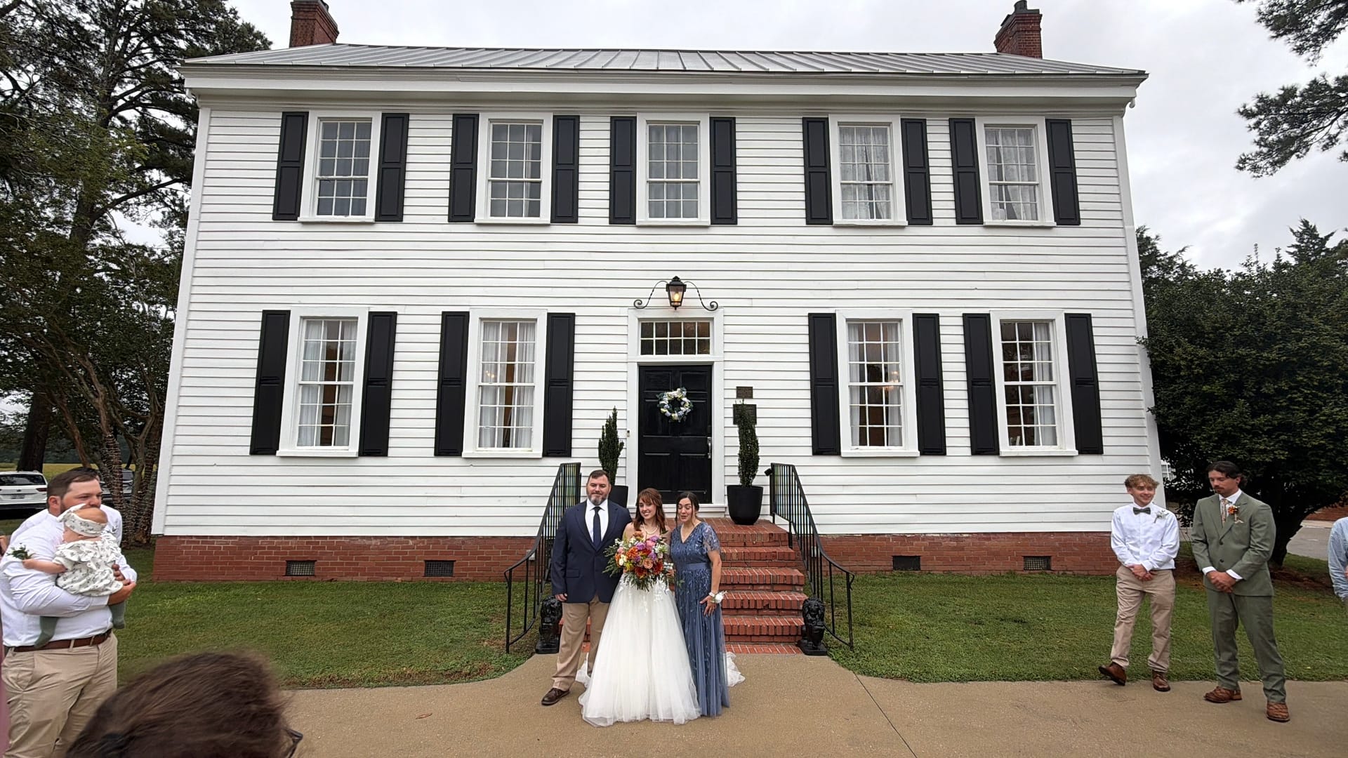Bride in a white tulle gown holding a colorful bouquet poses with two family members on the front steps of the 1821 Scarborough House white colonial manor with black shutters.