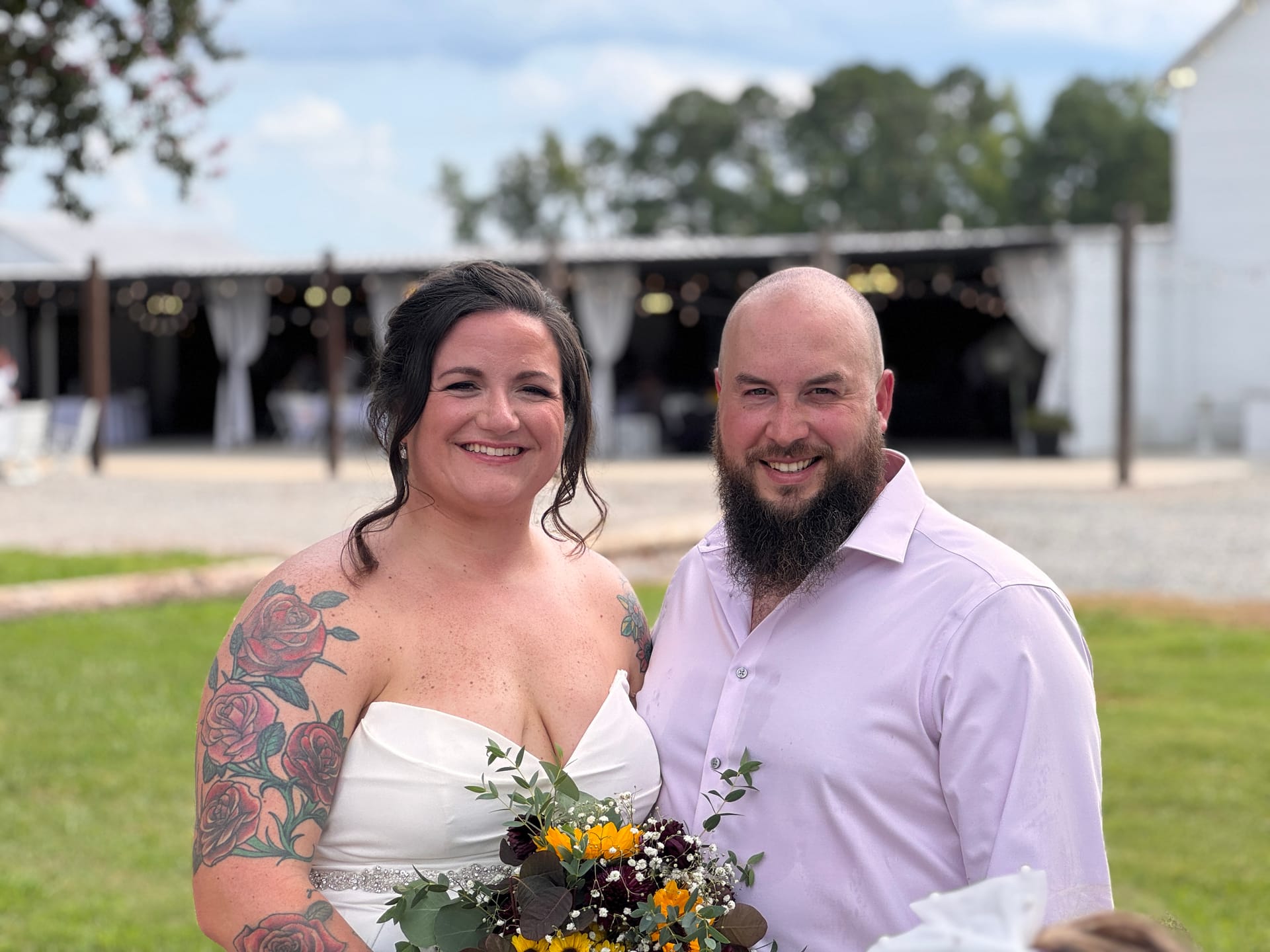 Smiling bride in a white strapless gown with rose sleeve tattoos holding a sunflower bouquet stands beside her bearded groom in a lavender dress shirt on the ceremony lawn at Scarborough House.