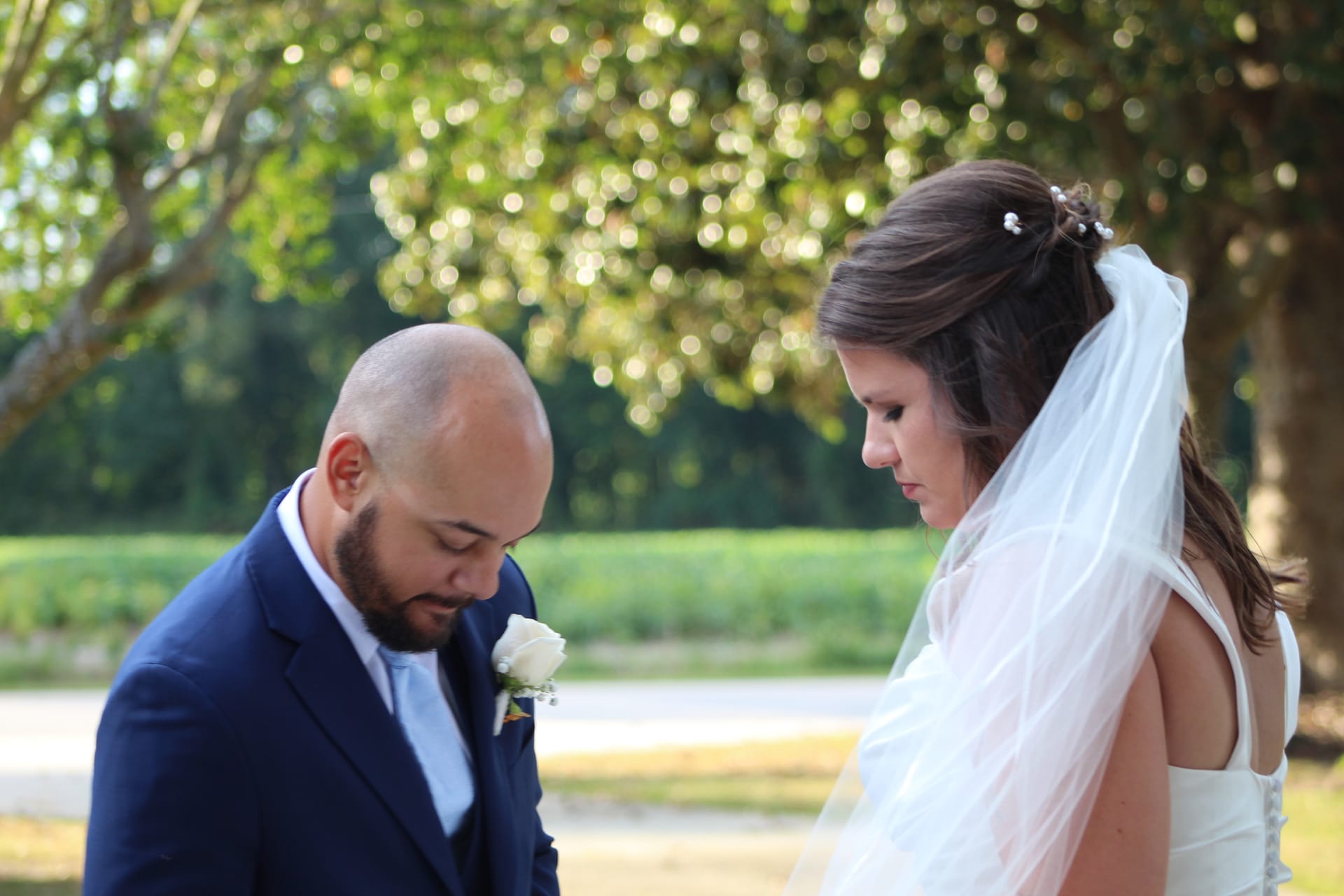 Bride in white gown and veil and groom in navy suit bow their heads together in a quiet reflective moment surrounded by lush green trees at Scarborough House.