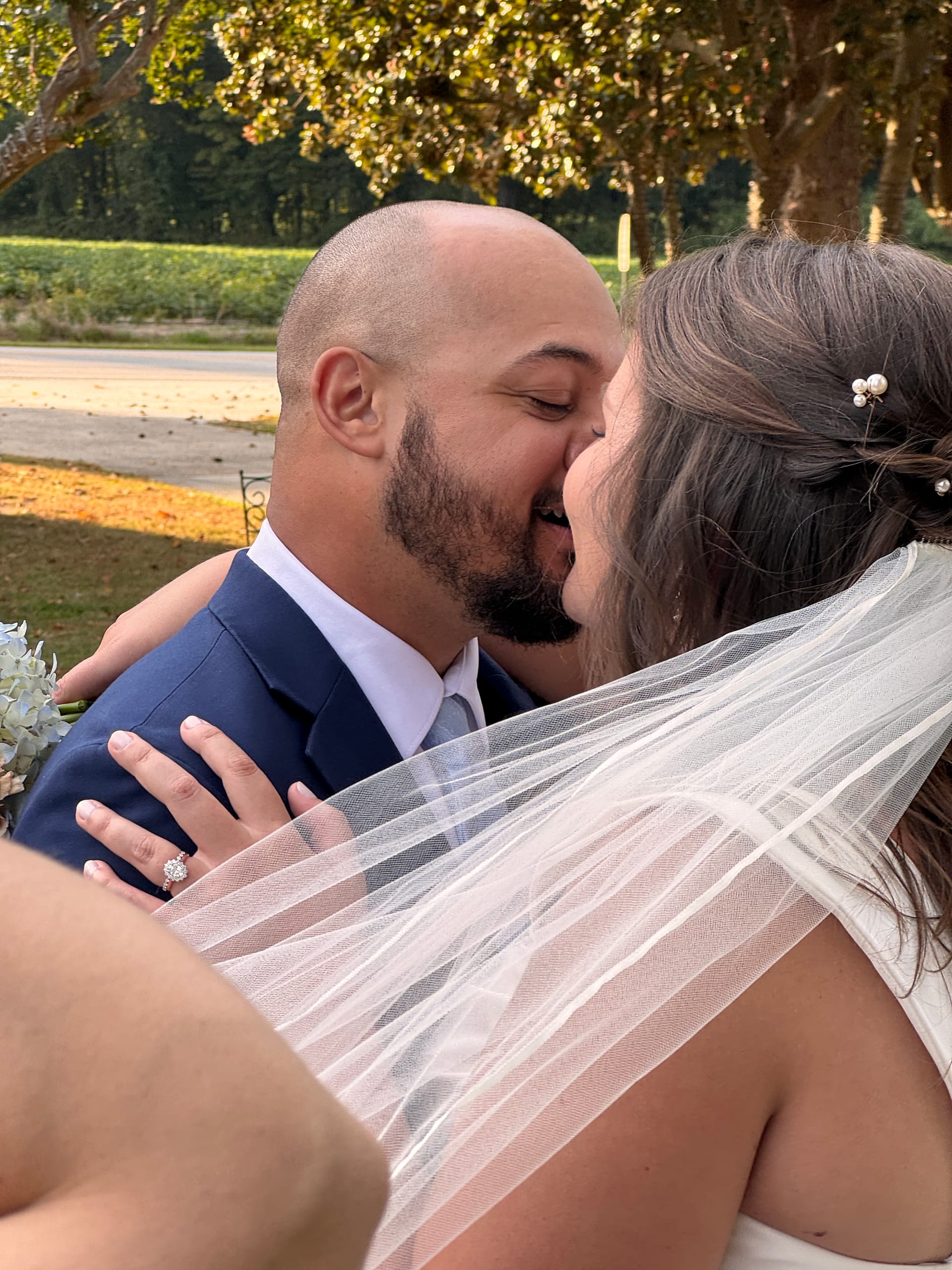 Bride and groom share their first kiss beneath golden-lit oak trees at Scarborough House estate, her white veil and pearl hair pins glowing in the warm afternoon light.