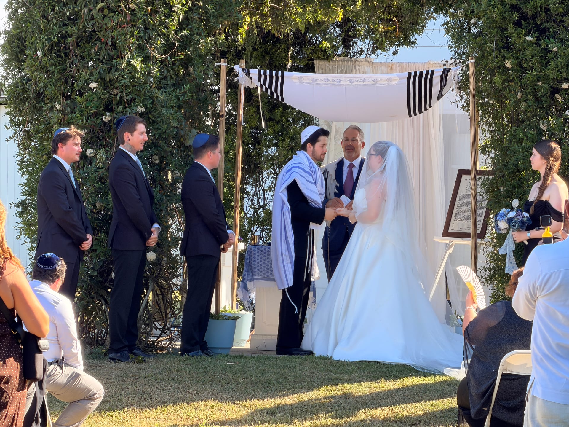 A bride and groom exchange vows beneath a white draped chuppah adorned with a tallit on the ceremony lawn surrounded by lush greenery at Scarborough House.
