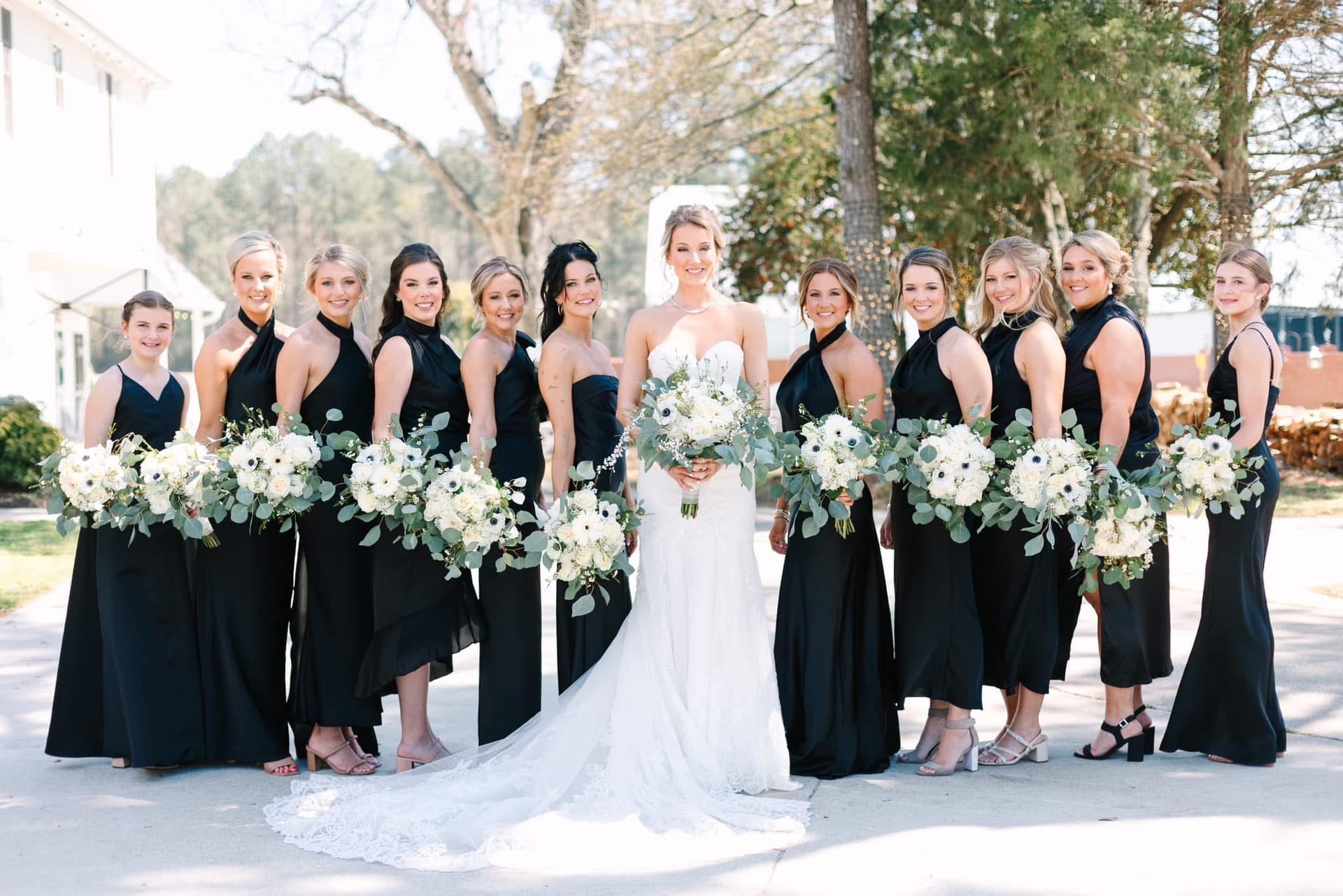 Bride in a white strapless gown stands centered among eleven bridesmaids in black halter-neck dresses holding white and green bouquets in front of a historic estate venue.