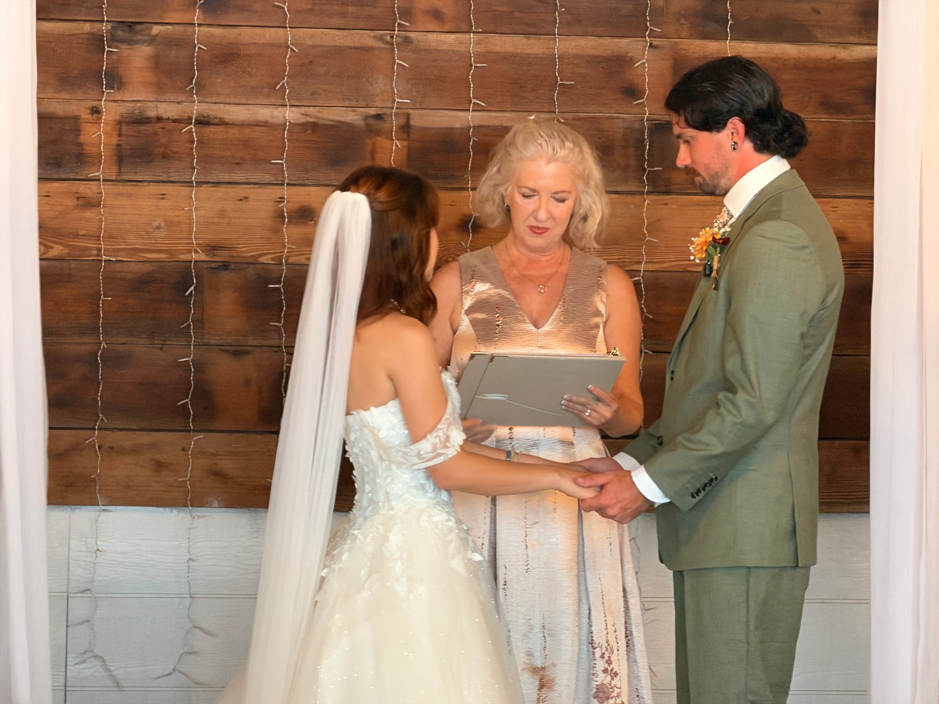 Bride and groom hold hands during their indoor wedding ceremony as the officiant reads from a gray book in front of a rustic wood plank wall adorned with string lights.