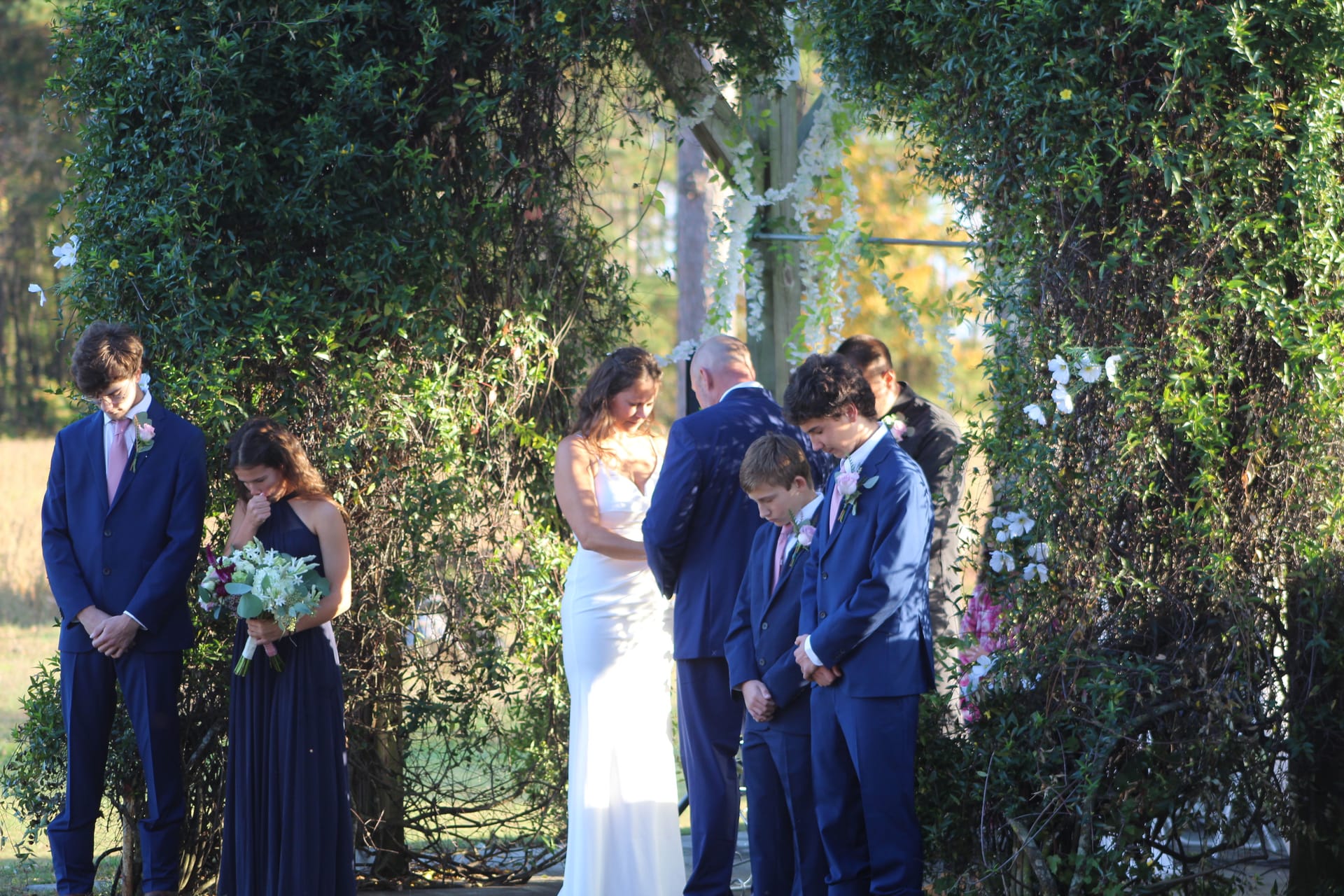 Wedding party bows heads in prayer beneath a lush floral arch during an outdoor ceremony at Scarborough House historic estate venue.