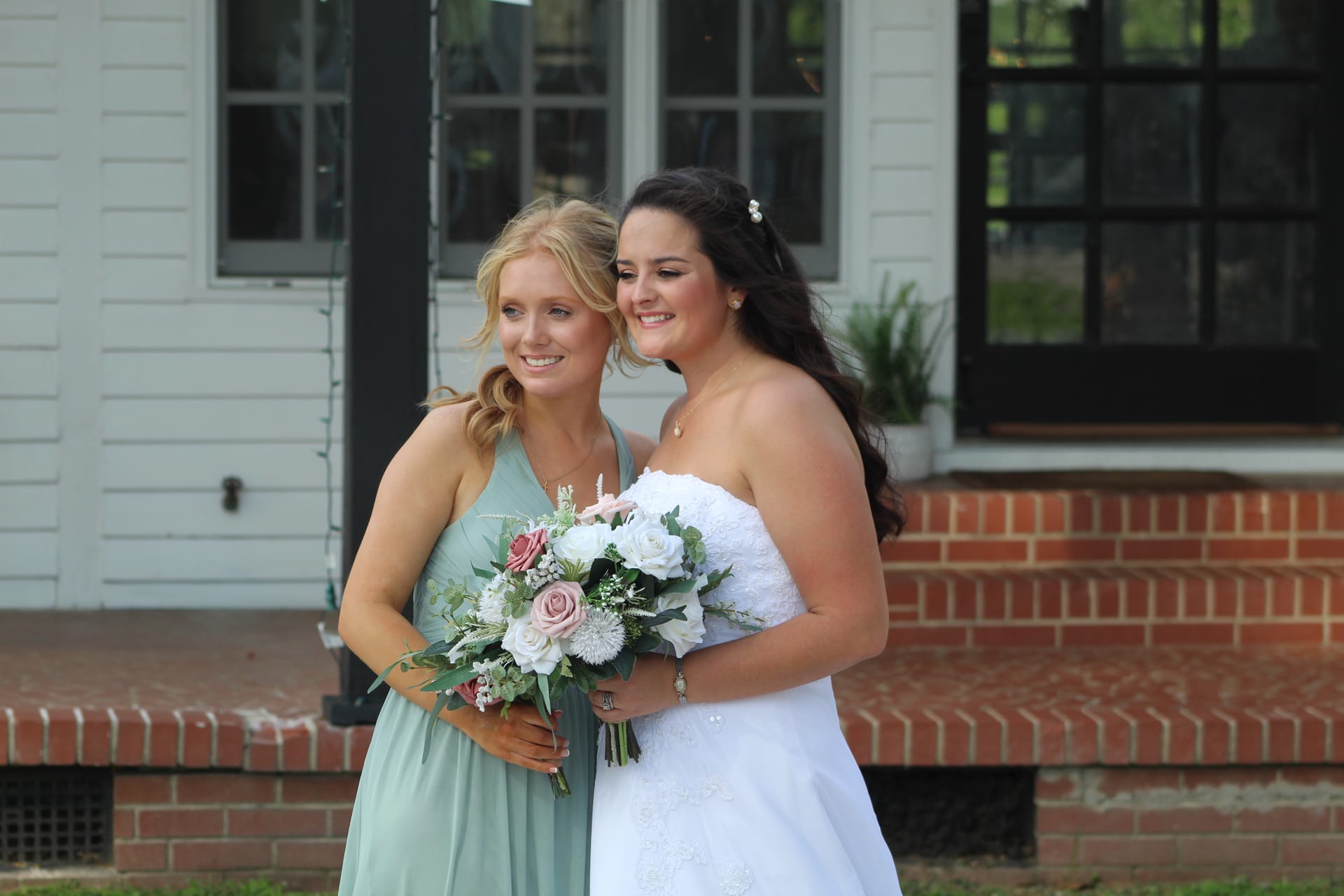 Bride in a strapless white lace gown and bridesmaid in sage green holding a blush and white floral bouquet smile together in front of the historic 1821 Scarborough House manor.