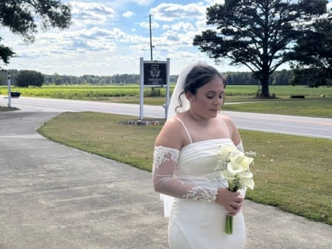Bride in a white off-shoulder lace gown holding white calla lilies stands near the Scarborough House entrance sign with green farmland fields in the background.