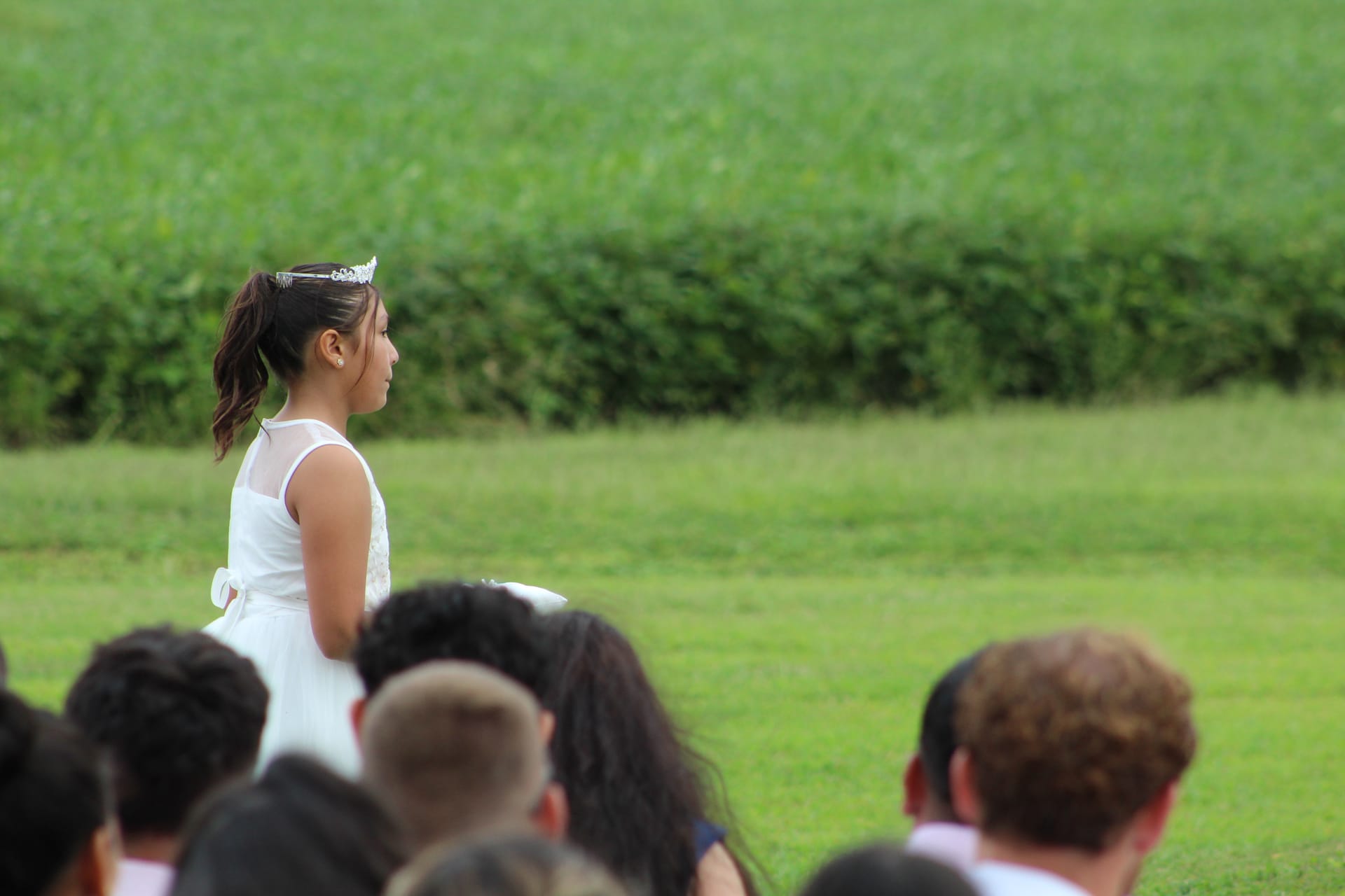 A young flower girl in a white dress and tiara stands on the ceremony lawn at Scarborough House with expansive green fields in the background.