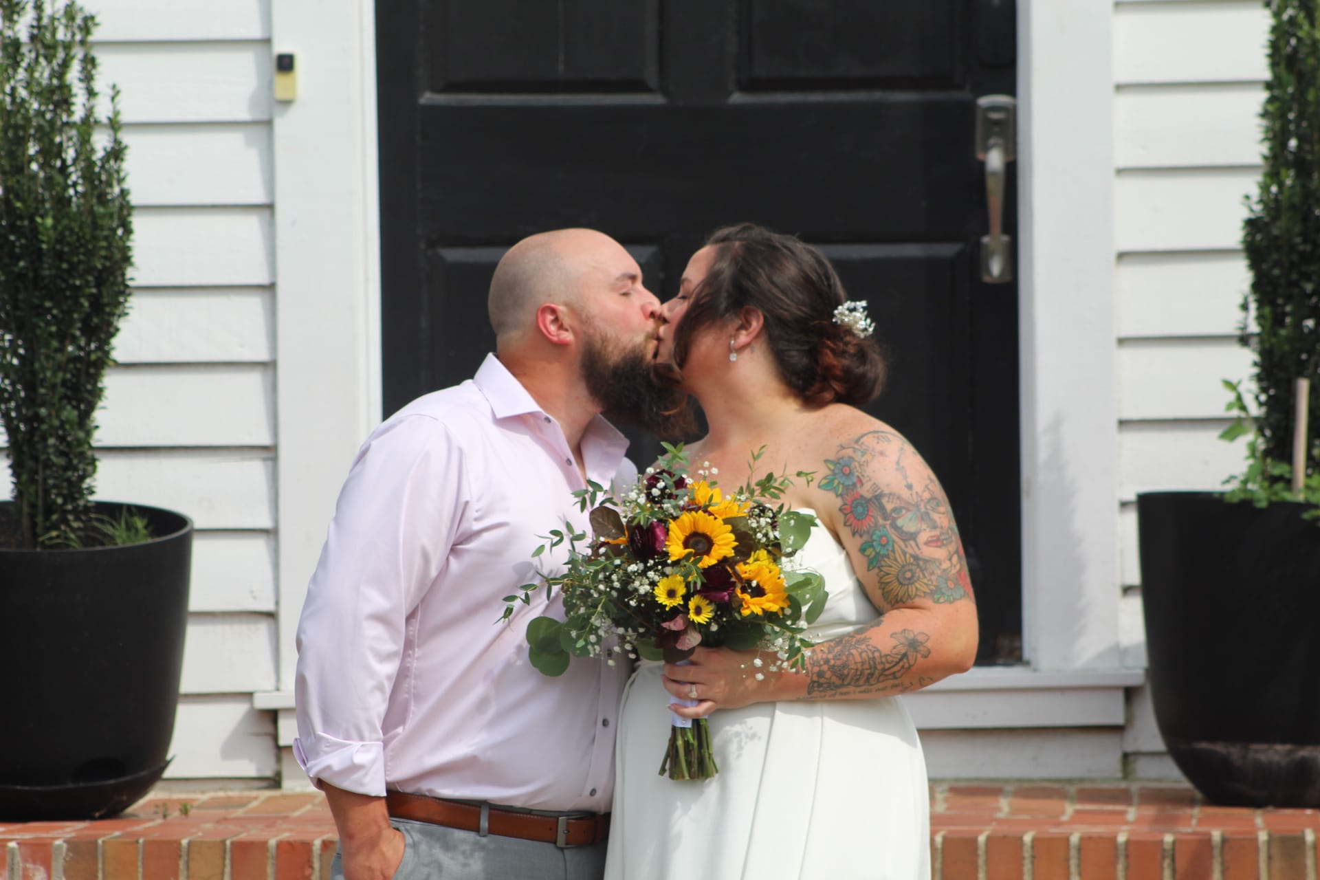Bride and groom share a romantic kiss on the front steps of the 1821 Scarborough House historic estate, bride holding a vibrant sunflower and greenery bouquet.