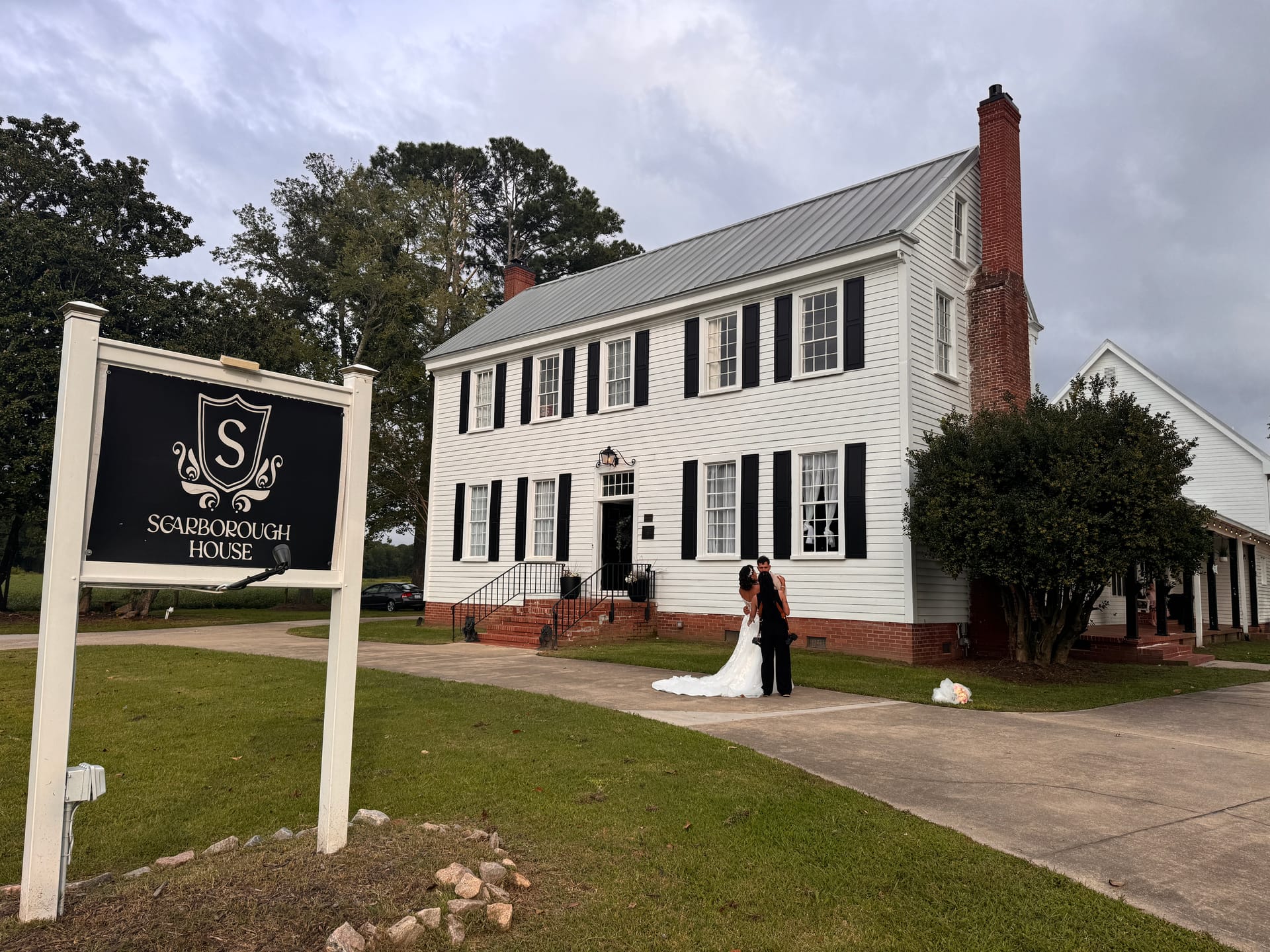 Bride and groom pose together in front of the historic white Scarborough House manor with its iconic venue sign in the foreground on an overcast day.