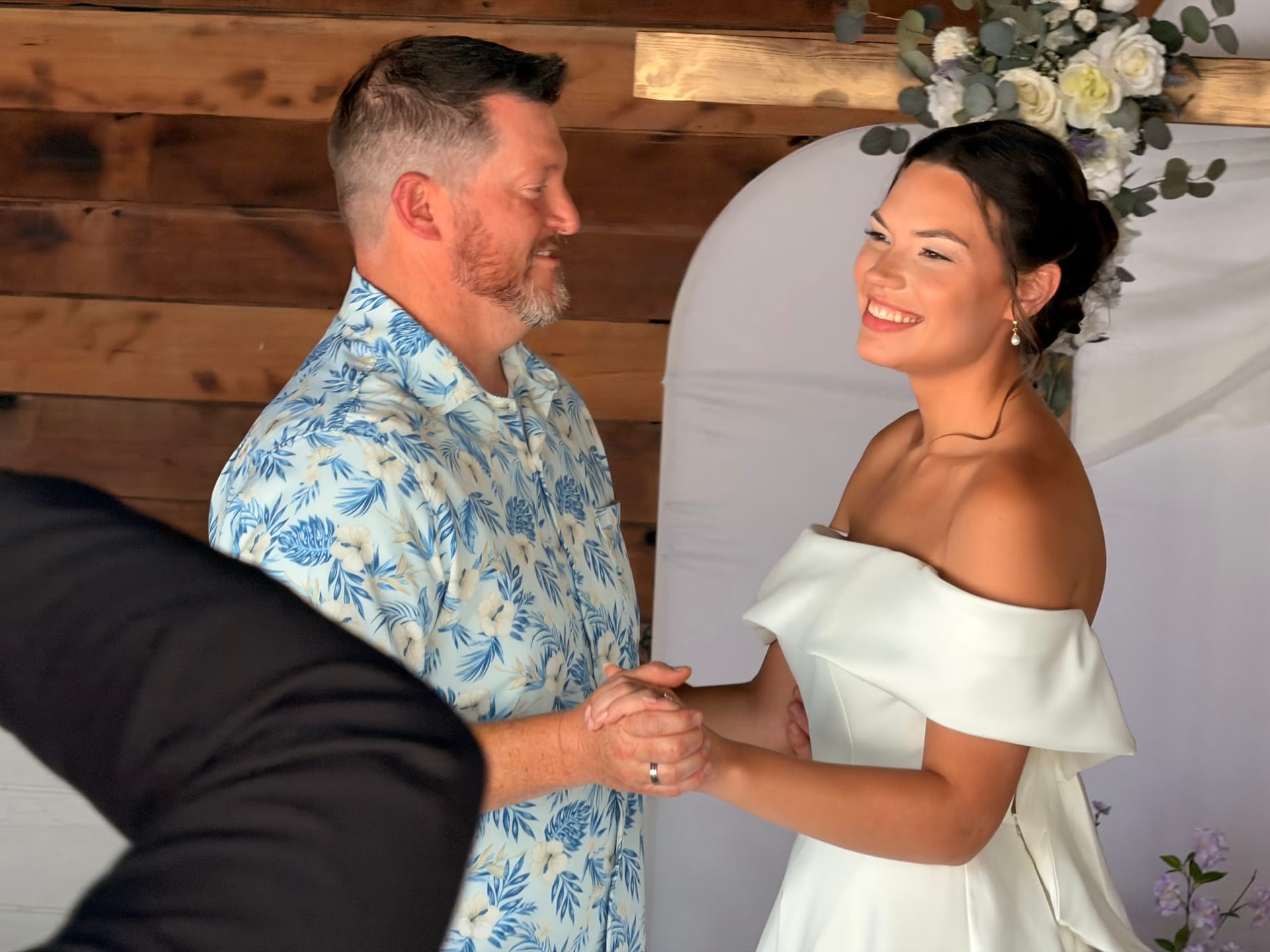A smiling bride in an off-shoulder white gown holds hands with the groom beneath a floral arch during their wedding ceremony at Scarborough House.