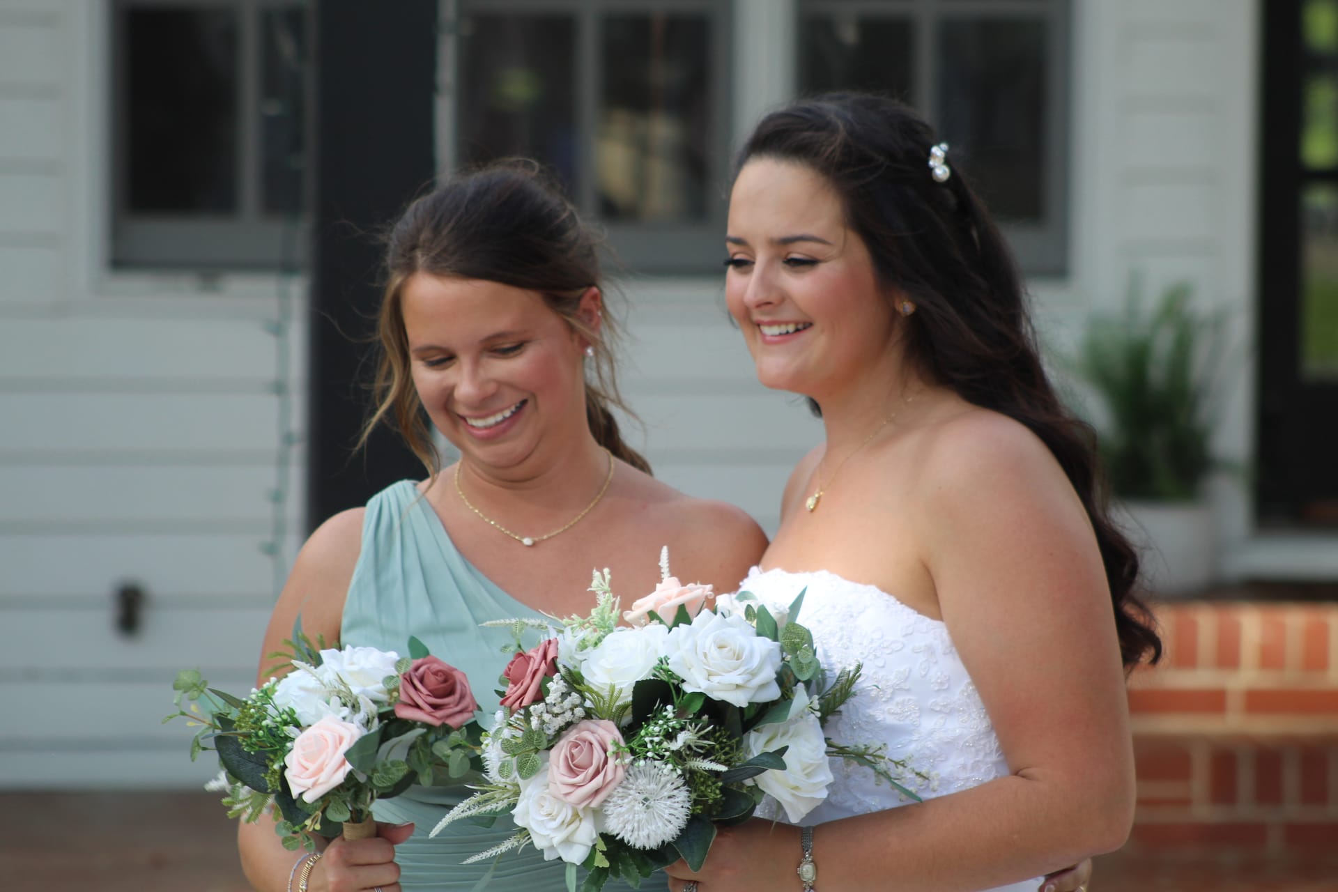 Bride in a strapless white lace gown and bridesmaid in a sage green dress share a joyful laugh while holding blush and white floral bouquets outside the historic Scarborough House manor.