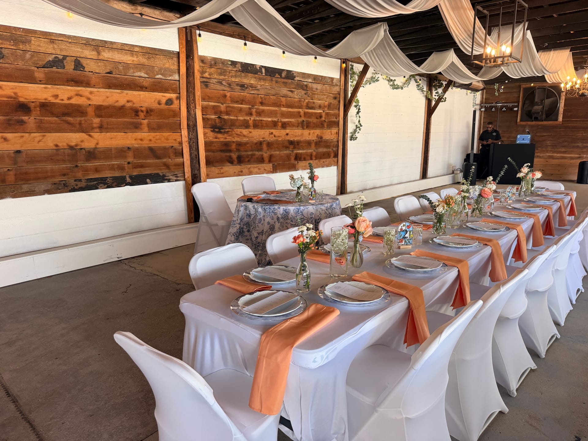 Elegantly set long reception table with white linens, coral satin napkins, and delicate floral centerpieces inside a rustic barn venue with draped fabric ceiling and string lights.