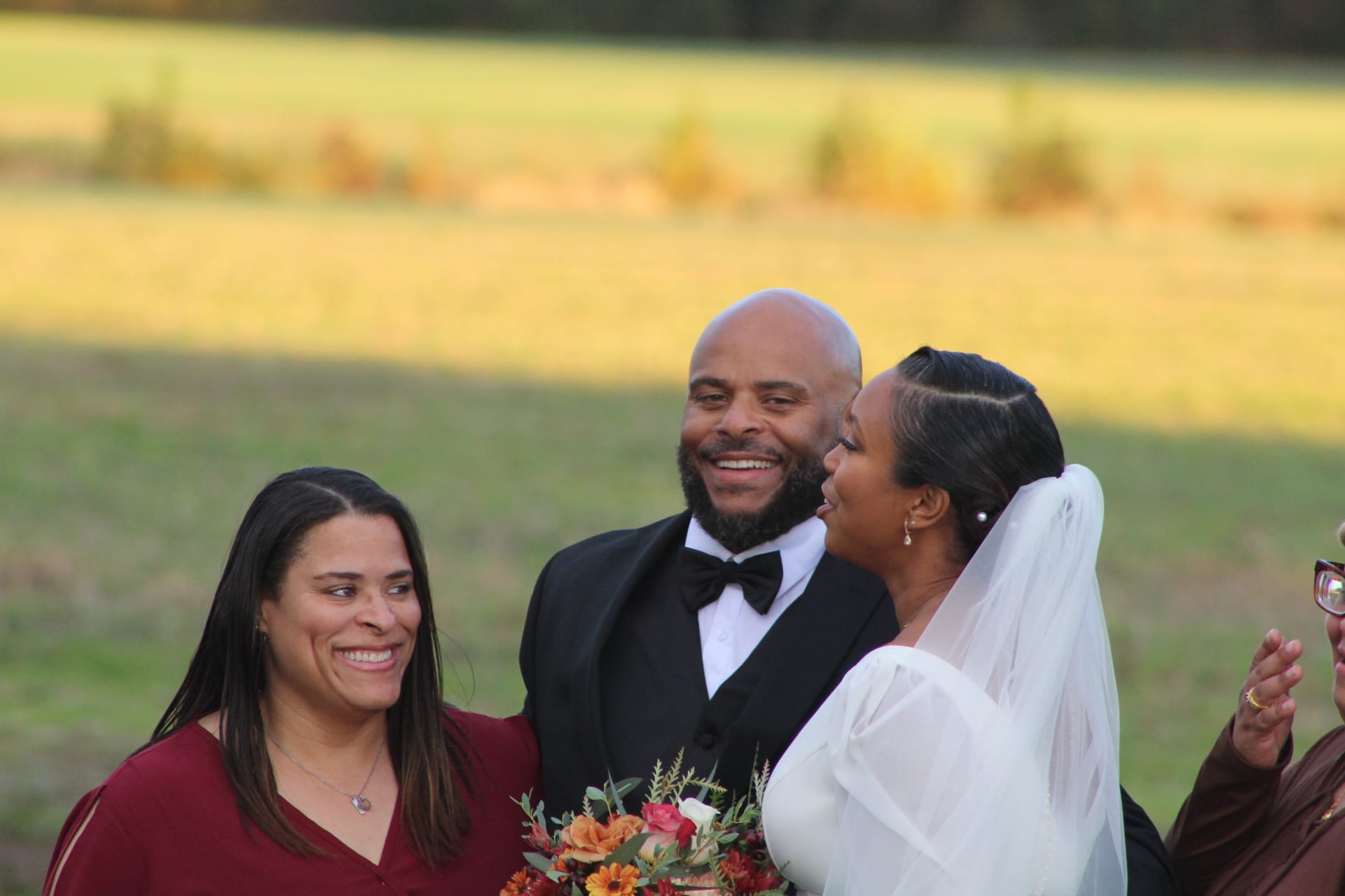 A smiling groom in a black tuxedo shares a joyful laugh with his bride in a white gown and veil and a bridesmaid in burgundy against a sunlit farmland field backdrop at Scarborough House.