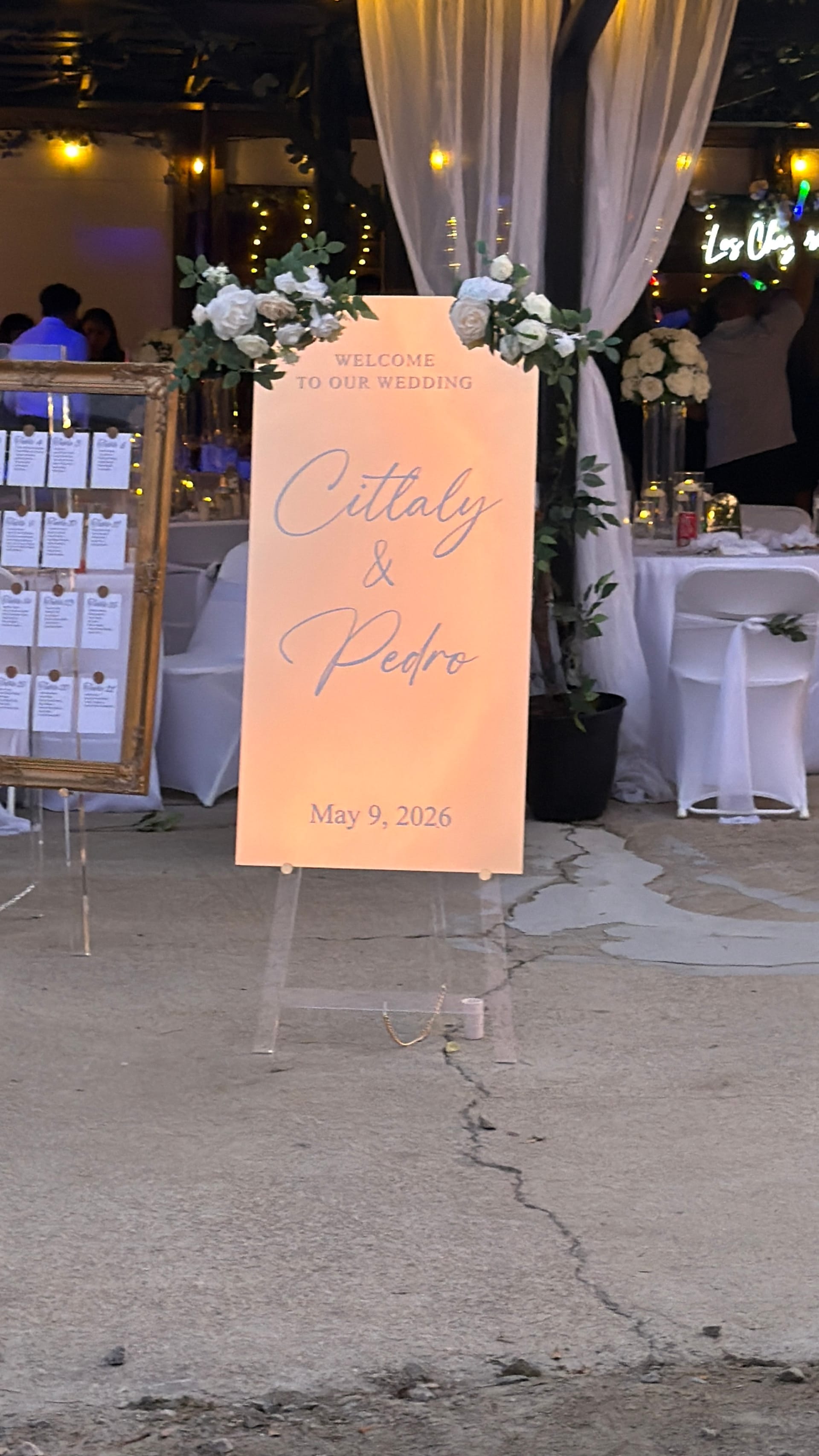 Peach acrylic wedding welcome sign for Cittaly and Pedro on May 9 2026 displayed on a clear easel adorned with white roses and greenery at a warmly lit reception venue.