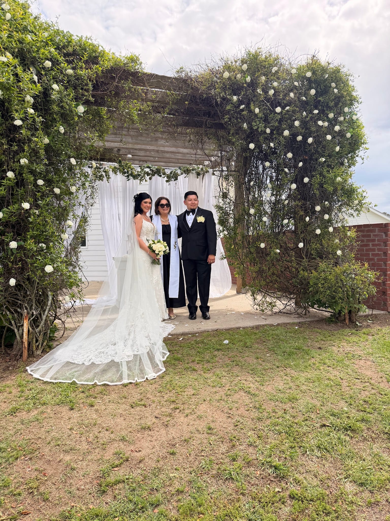 Bride in a lace ball gown and groom in a black tuxedo stand with their officiant beneath a lush green arbor adorned with white roses at Scarborough House.