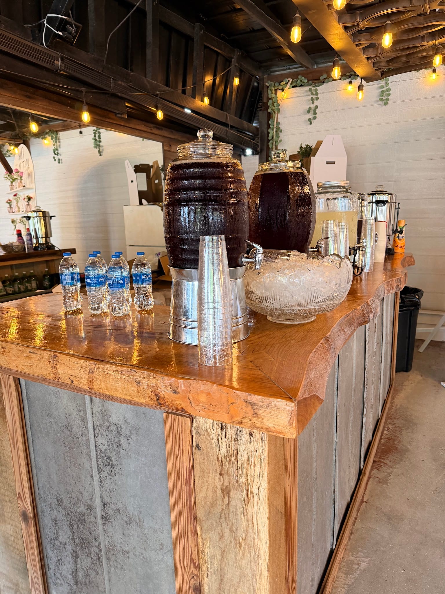 Rustic live-edge wooden bar at Scarborough House adorned with barrel drink dispensers, a glass lemonade jar, bottled water, and warm Edison string lights overhead.