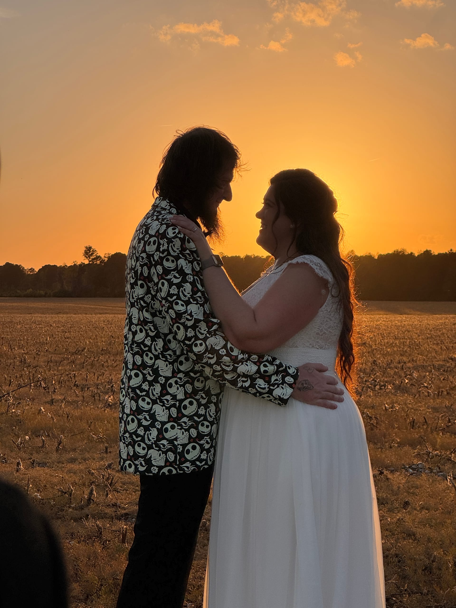 Bride in white lace gown and groom in a Nightmare Before Christmas print jacket embrace face-to-face in a harvested farmland field at Scarborough House during a vivid golden-hour sunset.