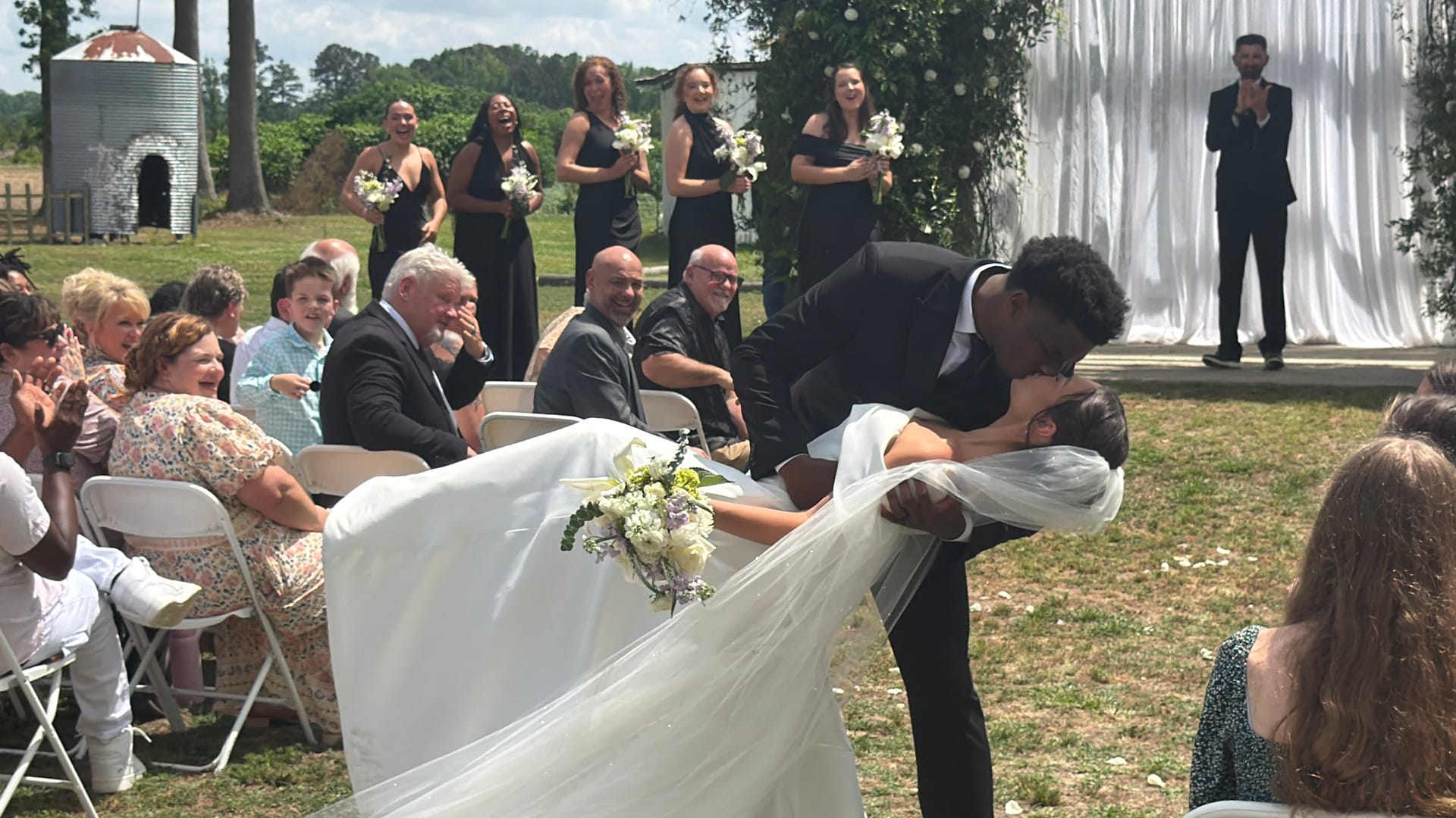 Groom dips and kisses bride in a dramatic mid-aisle moment surrounded by cheering guests and bridesmaids in black dresses at an outdoor farm estate wedding ceremony.